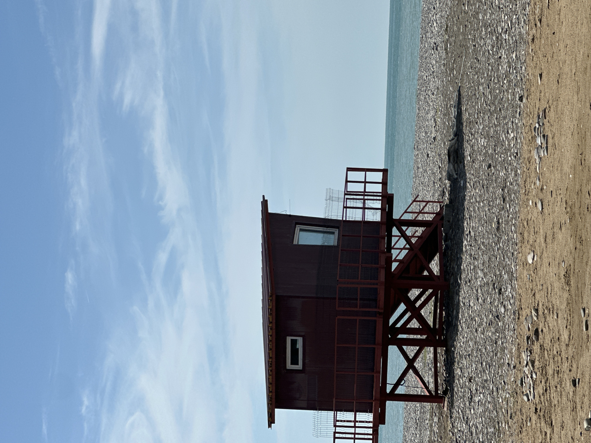 A lifeguard tower stands on a pebble and sand beach. The tower is dark red-brown in color, with a dark red base. It has two visible windows. The beach is divided into a section of gray pebbles and a section of tan sand. The sky is light blue with thin, wispy clouds. The Black Sea is visible in the background. The location is likely near Batumi, Georgia, given the type of beach, and the architecture of the building.