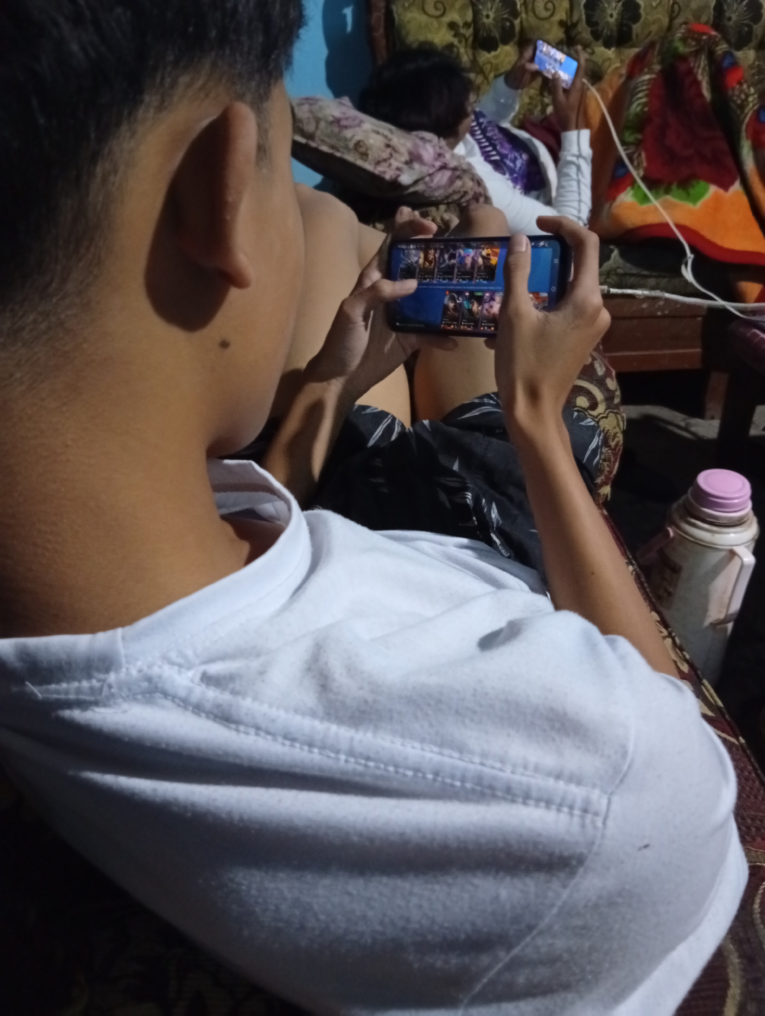 A medium shot from behind shows a young person in a white t-shirt playing a mobile game. The person is holding the phone horizontally with both hands. The phone screen displays colorful icons and what appears to be a game interface. In the background, another person is visible, also looking at a mobile phone. They are lying down on a patterned surface, possibly a bed or couch, covered with blankets. A patterned fabric with floral designs is visible. A thermos with a pink lid is on the right side of the frame. The scene appears to be indoors, with a blue wall in the upper left corner. The lighting is dim. This scene is likely located in a residential area in Mriyan, Indonesia.