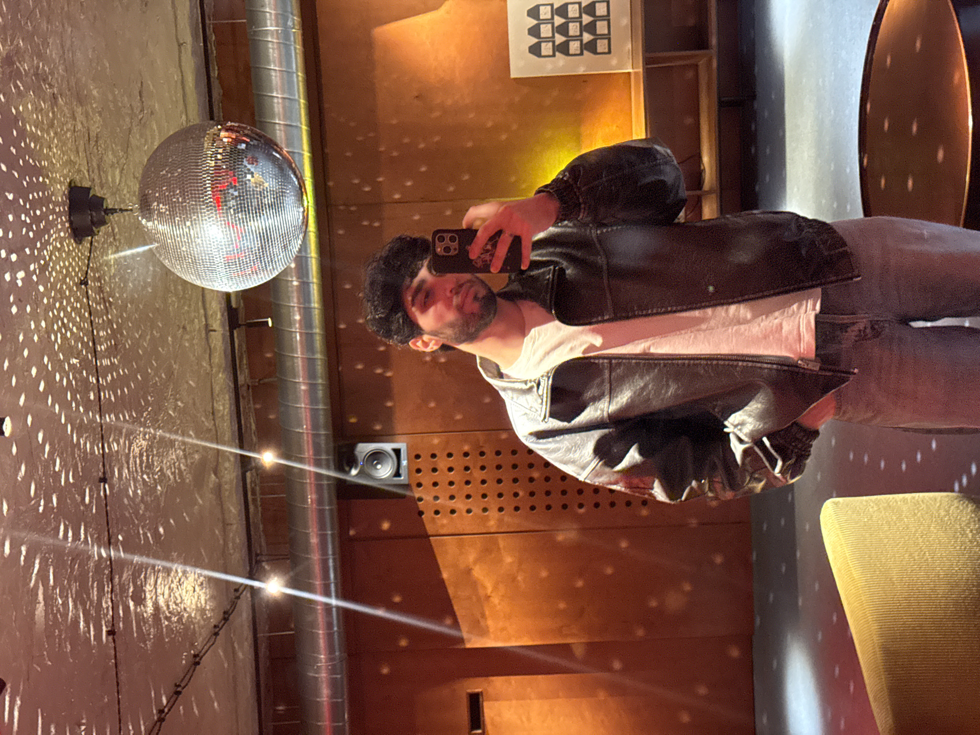A man with dark hair and a beard is holding a smartphone to take a selfie in a room illuminated by the reflections of a disco ball. He is wearing a black leather jacket over a white shirt and gray jeans. The room has a textured wall with a metallic pipe and a speaker mounted on it. In the background, there are shelves and a seating area. The location is possibly in Yerevan, Armenia, although this cannot be confirmed from the image.
