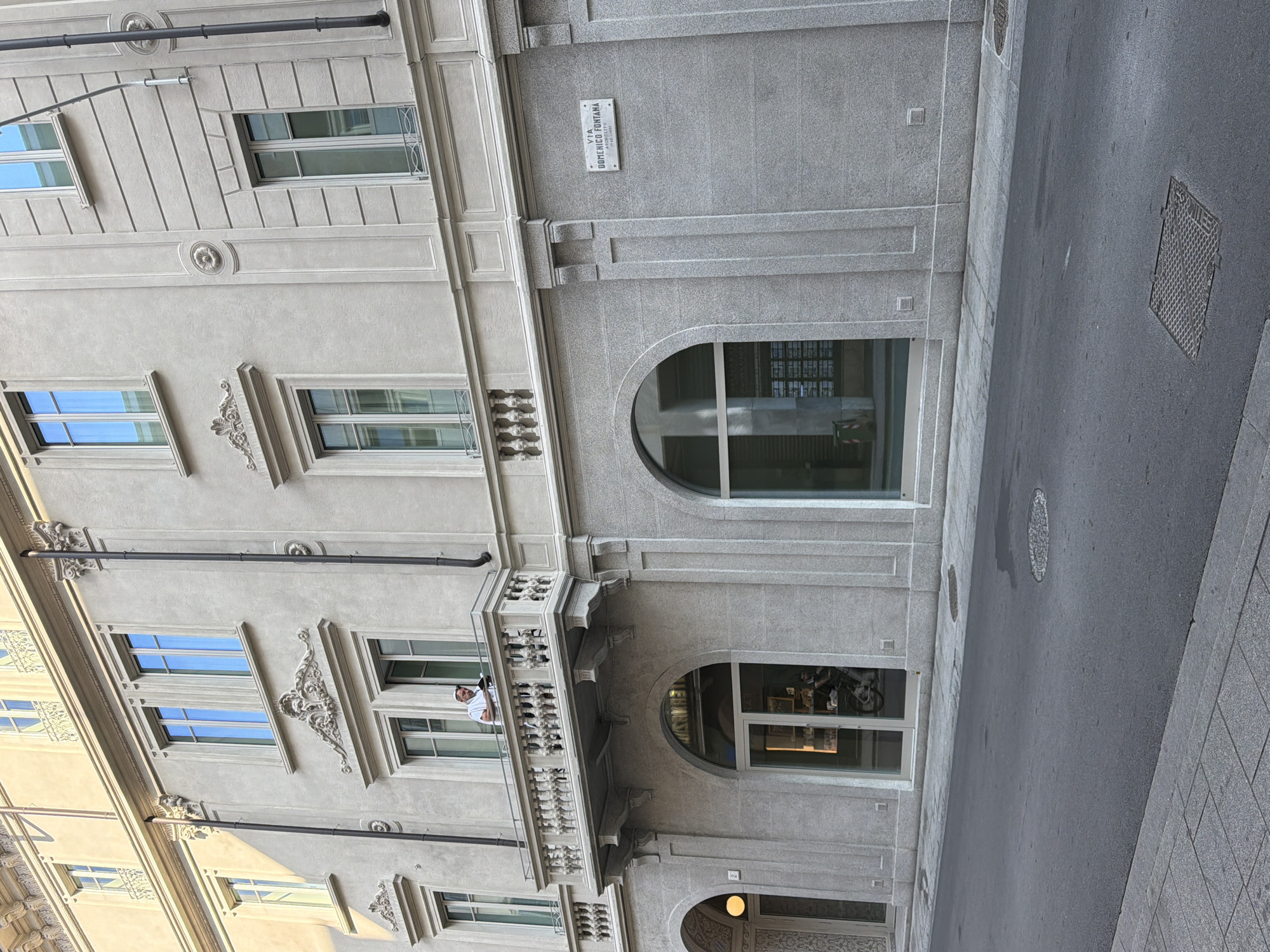 Here is a description of the image:
The image shows the architecture of a building located on Via Domenico Fontana in Como, Italy. The building has a stone facade with windows and doorways, some arched. There's a balcony where a man stands. A street runs alongside the building. A sign bearing the street name "Via Domenico Fontana" is visible on the building's facade.