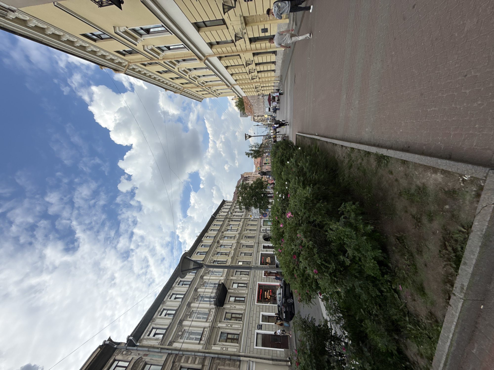 The image presents a daytime city street scene, captured from a low, upward-looking angle. Two multi-story buildings define the street. The building on the left (when correctly oriented) features a light, possibly cream or pale yellow facade with decorative cornices and numerous windows. The building on the right is similarly multi-storied, with a gray-beige facade and many windows.
A paved pedestrian area runs along the right side of the street. Several individuals are present: two people are walking in the foreground, one in light attire and another in darker clothes with a backpack. Further down the street, more people are visible, some seated at outdoor tables, suggesting cafes or restaurants. A line of green bushes, some bearing small pink flowers, separates the paved area from what appears to be a narrower street or pathway. A black car is parked near the right-hand building.
The activity taking place is typical urban life, with people walking, standing, and dining outdoors. The time of day is daytime, with clear, bright lighting. The weather is partly cloudy, featuring white cumulus clouds against a blue sky, indicative of a pleasant day. Overhead wires are visible stretching across the street.
Notable visible text includes signs on the right-hand building. One prominent sign, partially visible and in Cyrillic script, reads "КРАФТОВАЯ" (CRAFT) and another below it says "БУРГЕРНАЯ" (BURGER JOINT), strongly suggesting the location is in a Russian-speaking country. The architecture and street design imply an older European urban setting.