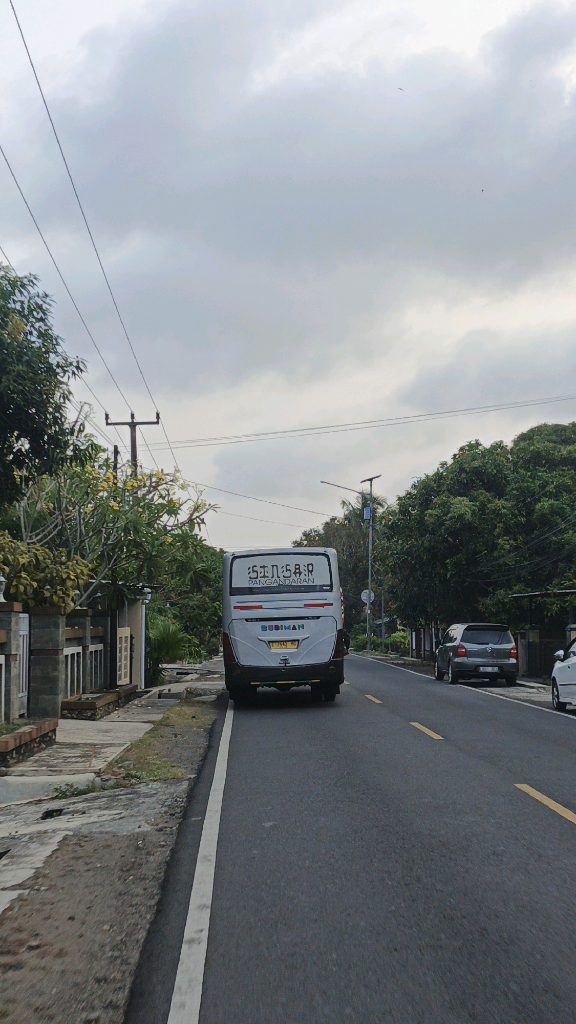 An overcast sky looms over a paved road in Karangjaladri, Indonesia. A white bus with the text "5 I N 5 B R" and "PANGANDARAN" is in the center of the frame, facing away from the camera. The bus also has the name "BUDIMAN" printed on its rear, along with a license plate that reads "E 7542 MG". To the left of the road, a sidewalk is bordered by a wall and lush green trees. On the right side of the road, a silver car is parked. The road itself has a white line marking the edge and yellow dashed lines in the center. Power lines and poles are visible against the cloudy sky.