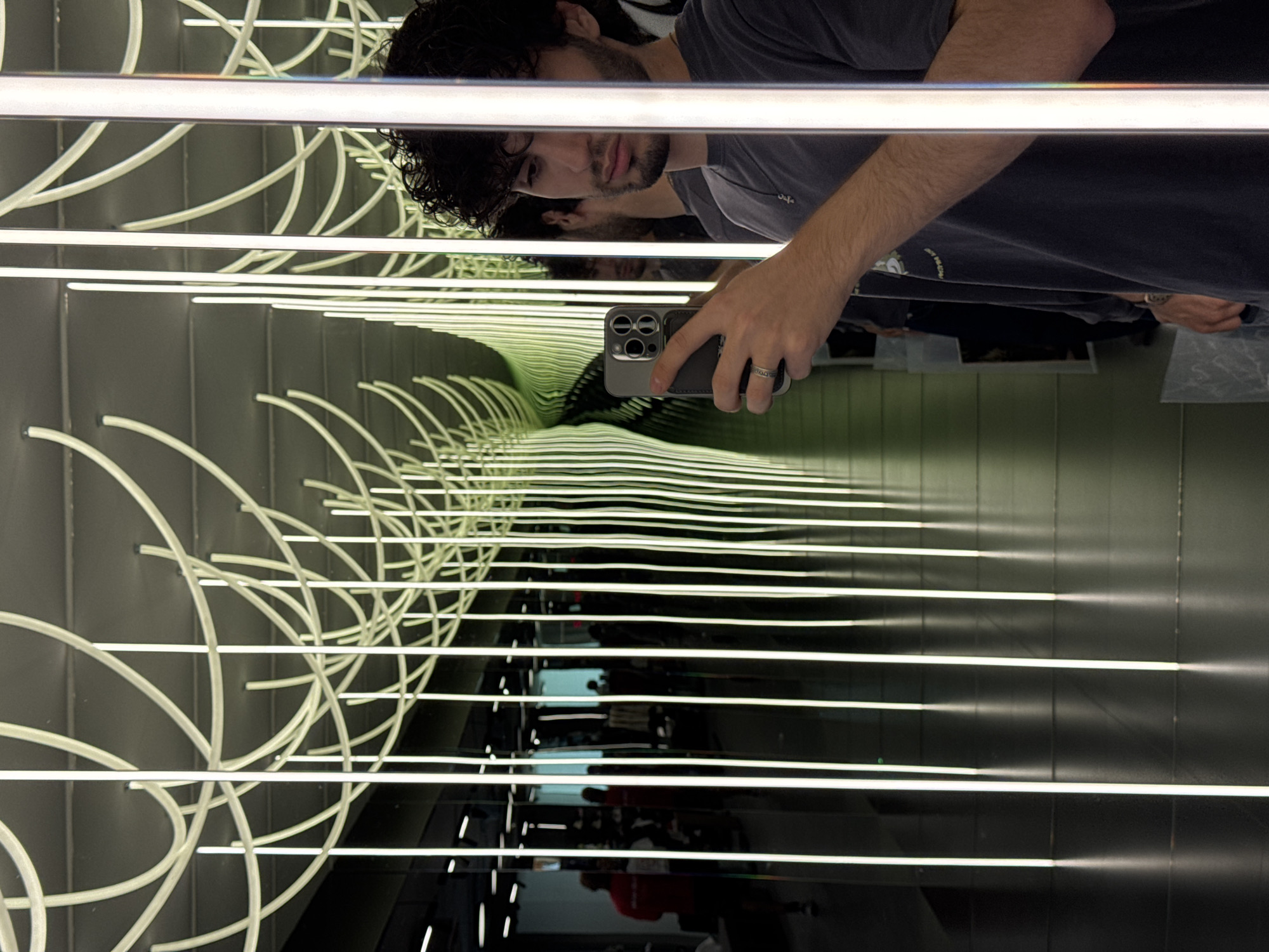 This image is a selfie taken in a room with mirrored walls and an infinity mirror effect created by strategically placed vertical LED lights and curved ceiling lights. The room appears to be an indoor retail space, possibly a store, with merchandise visible in the background reflections. The person taking the photo, a young man with curly dark hair, is wearing a dark t-shirt and a ring on his left hand. He is holding a smartphone in his right hand. The floor is dark and reflective, mirroring the lights and the surrounding environment. The perspective of the image suggests the photographer is standing near the entrance of a section of the store, perhaps a fitting room or a display area designed for an immersive experience. The location of this scene is the "Mirror Maze" at the Yerevan Mall in Yerevan, Armenia.