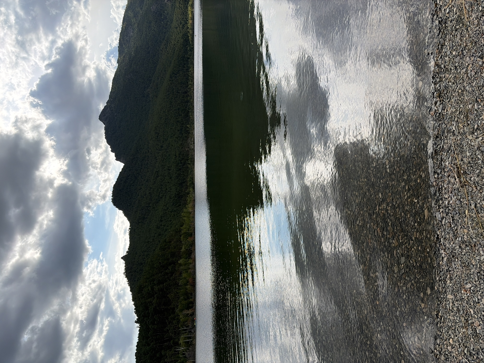 The image depicts a serene natural landscape with a body of water in the foreground, likely a lake, reflecting a cloudy sky and the silhouette of a forested mountain range. The shoreline is composed of small, gray and brown pebbles. The water is calm with gentle ripples, showing clear reflections of the sky and distant trees. The sky is dramatic, with large, dark clouds interspersed with patches of lighter sky, suggesting an overcast or partly cloudy day. The mountain range is densely covered in dark green coniferous trees. There are no people visible in the immediate foreground or on the water. The scene is a natural outdoor environment.
