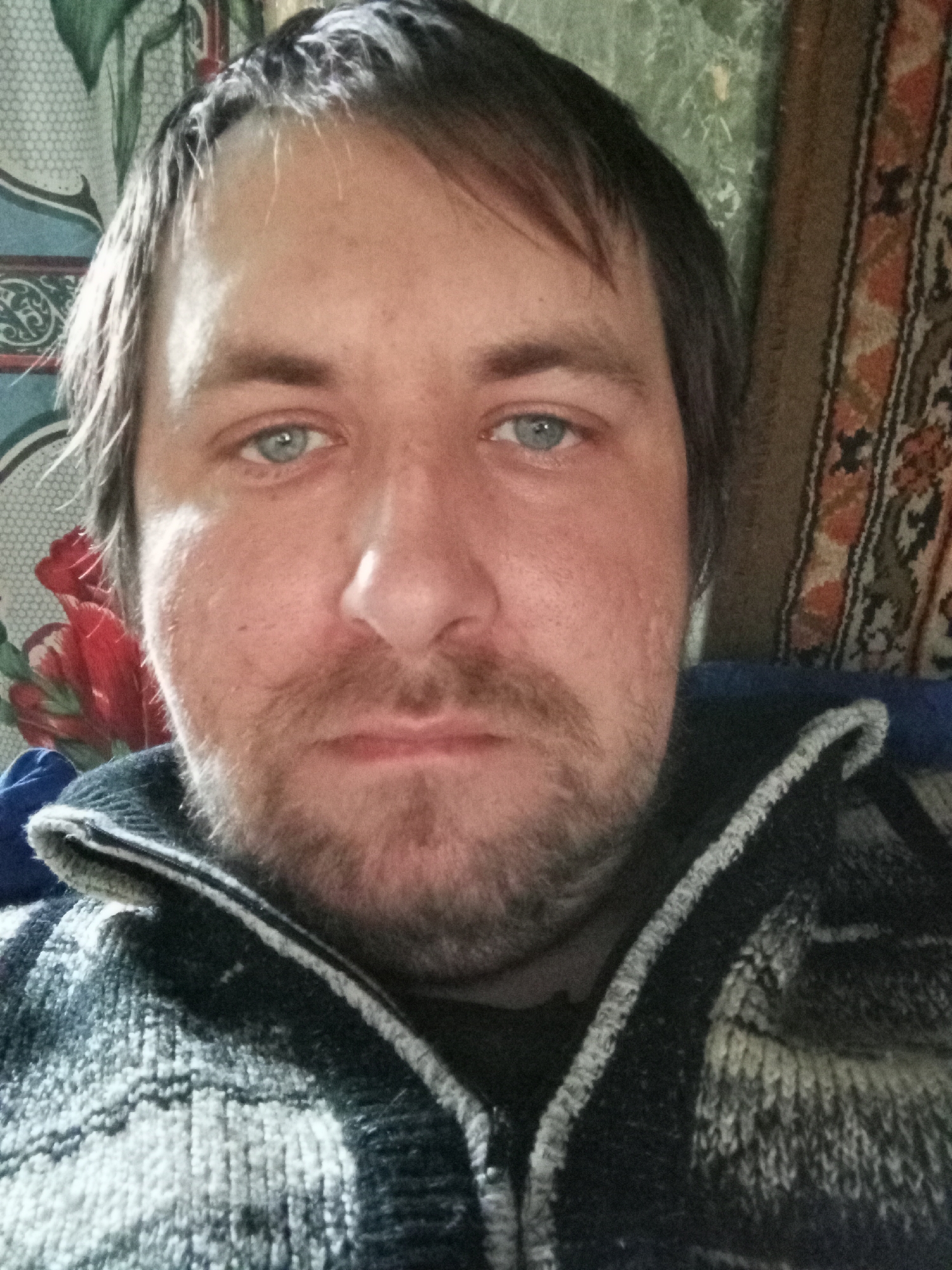 A self-portrait of a Caucasian male with fair skin, blue eyes, and dark brown hair that is partially receding at the hairline. He has a full, untrimmed beard and mustache. He is wearing a knitted sweater with a zipper, likely in a casual setting. The background is cluttered with floral-patterned and woven tapestry. There is no information in the image to determine the location is Parabel, Russia.