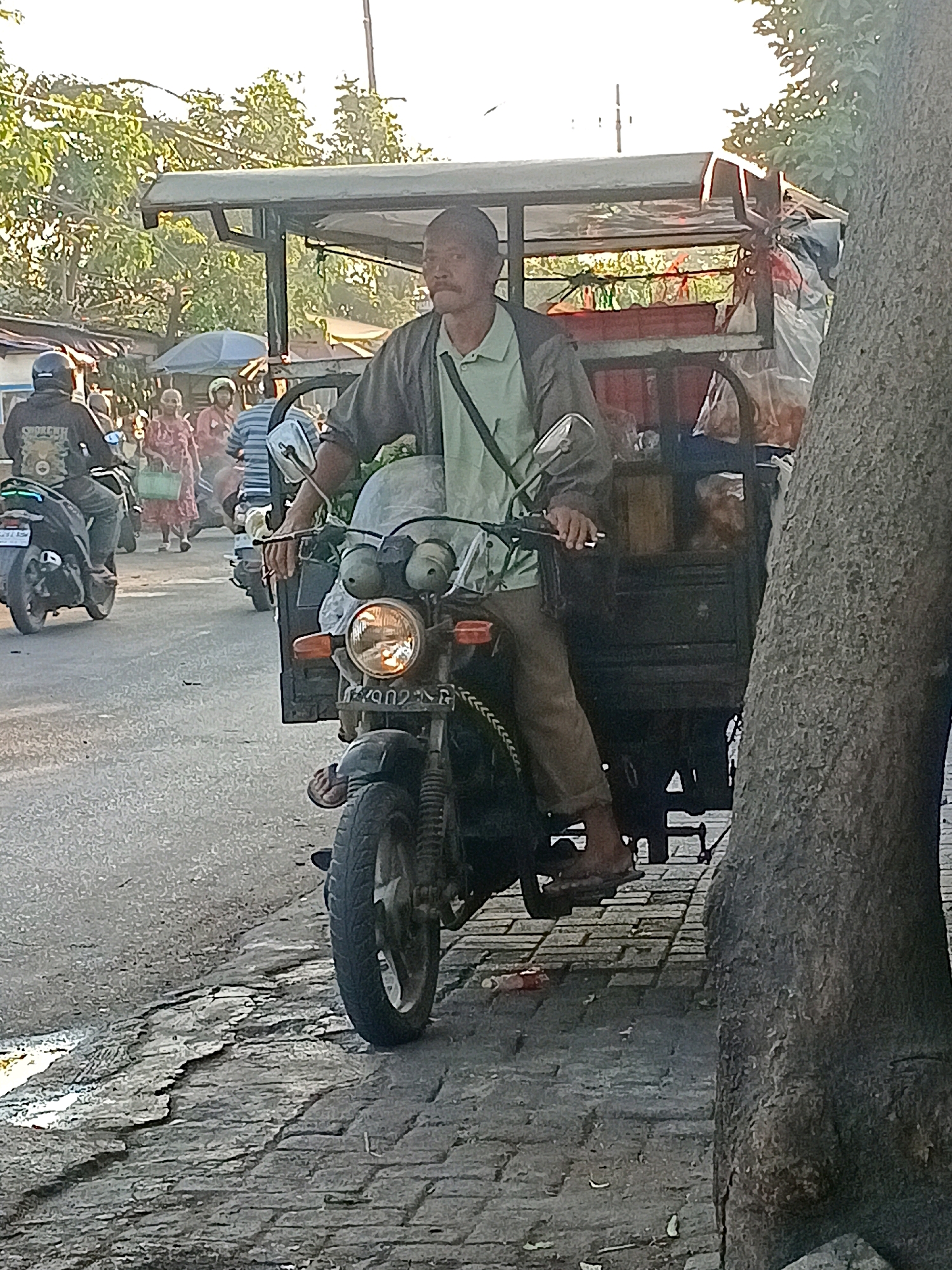 A medium-shot frontal view captures a man operating a three-wheeled motorcycle with a canopy and cargo area, parked on a cracked paved surface adjacent to a dirt road. The man, wearing a light green collared shirt and dark pants, is seated on the motorcycle, holding the handlebars. The motorcycle has a prominent headlight and an orange turn signal. The cargo area behind him is filled with various items, including plastic bags and what appears to be food products. In the background, several other motorcycles and people are visible, suggesting a bustling street scene in Wonocolo, Indonesia. The environment is illuminated by sunlight, with trees and buildings forming the backdrop. A large tree trunk is prominently positioned on the right side of the frame.