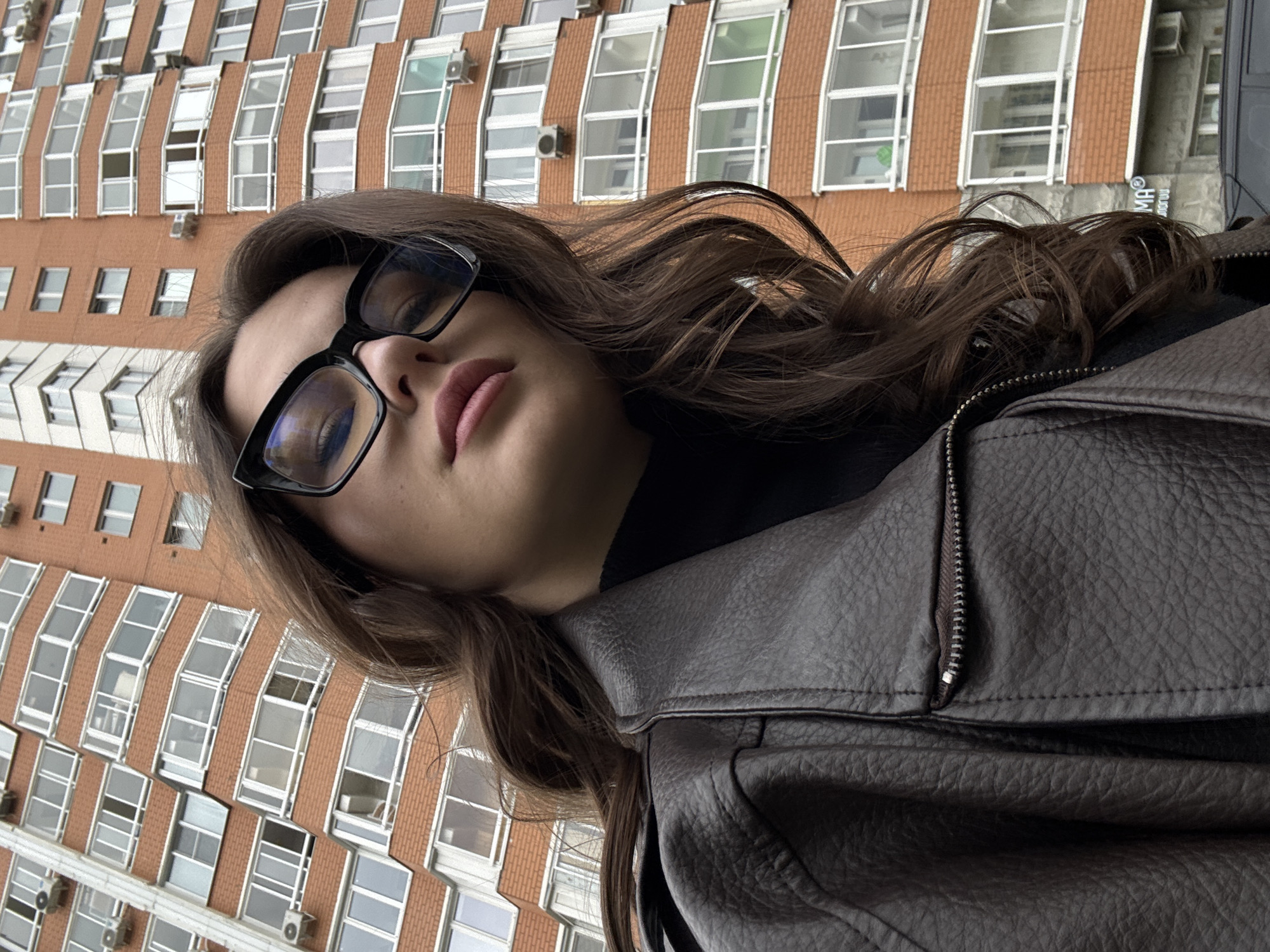 A low-angle shot captures a woman wearing glasses and a brown leather jacket. The backdrop features a tall brick building with rows of windows, suggestive of an urban setting. The location is in Vnukovo, Russia, based on the architectural style and urban landscape.