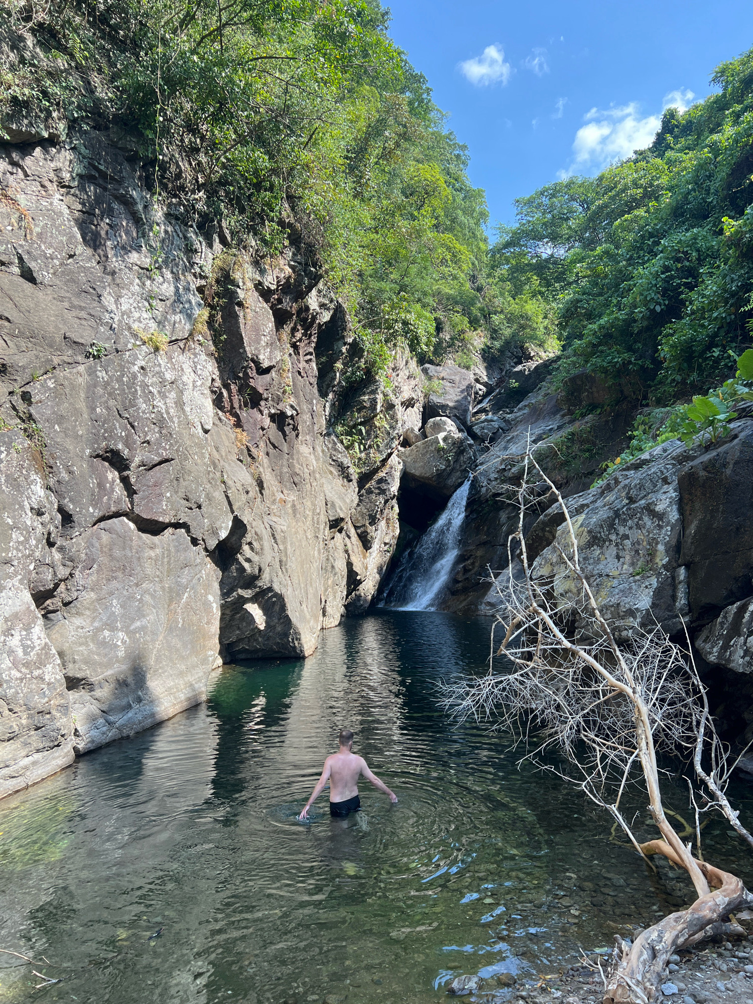 A man stands in a natural pool of water at the base of a waterfall, surrounded by lush green vegetation and large rock formations. The man, facing away from the camera, is partially submerged in the clear water, his arms outstretched. The waterfall cascades down a rocky cliff into the pool. The scene is illuminated by natural sunlight, casting shadows and highlighting the textures of the rocks and water. The image conveys a sense of natural beauty and tranquility. This location is likely a hidden gem within the natural landscapes surrounding the city of Da Nang, Vietnam, potentially in the Son Tra Peninsula or Ba Na Hills region, known for their waterfalls and dense forests.