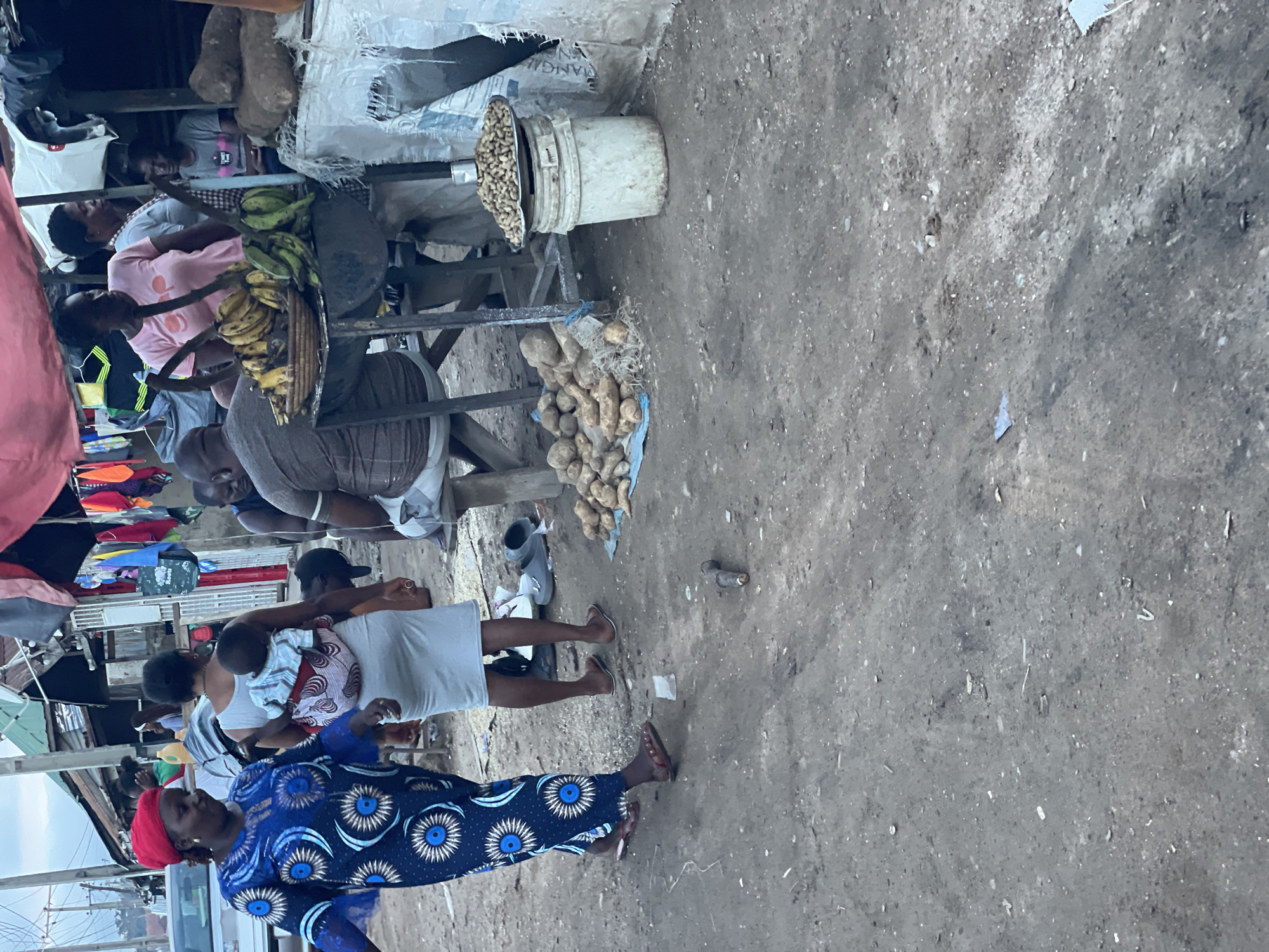 In an outdoor marketplace scene, likely in Abuja, Nigeria, several individuals are visible engaged in various activities. A woman in a blue and white patterned dress with a red headscarf is walking, facing left. To her right, another woman is carrying a baby on her back, wearing a white dress. In the background, several people are gathered around stalls. One man, wearing a grey t-shirt, is seated, and another person in a pink t-shirt is also visible. A variety of goods are displayed, including bananas on a table and a pile of yams or similar root vegetables on the ground. A bucket with a metal tray of peanuts sits beside a white sack. The ground is unpaved, and debris is scattered across it. The scene appears to be a bustling local market with a mix of commerce and daily life.
