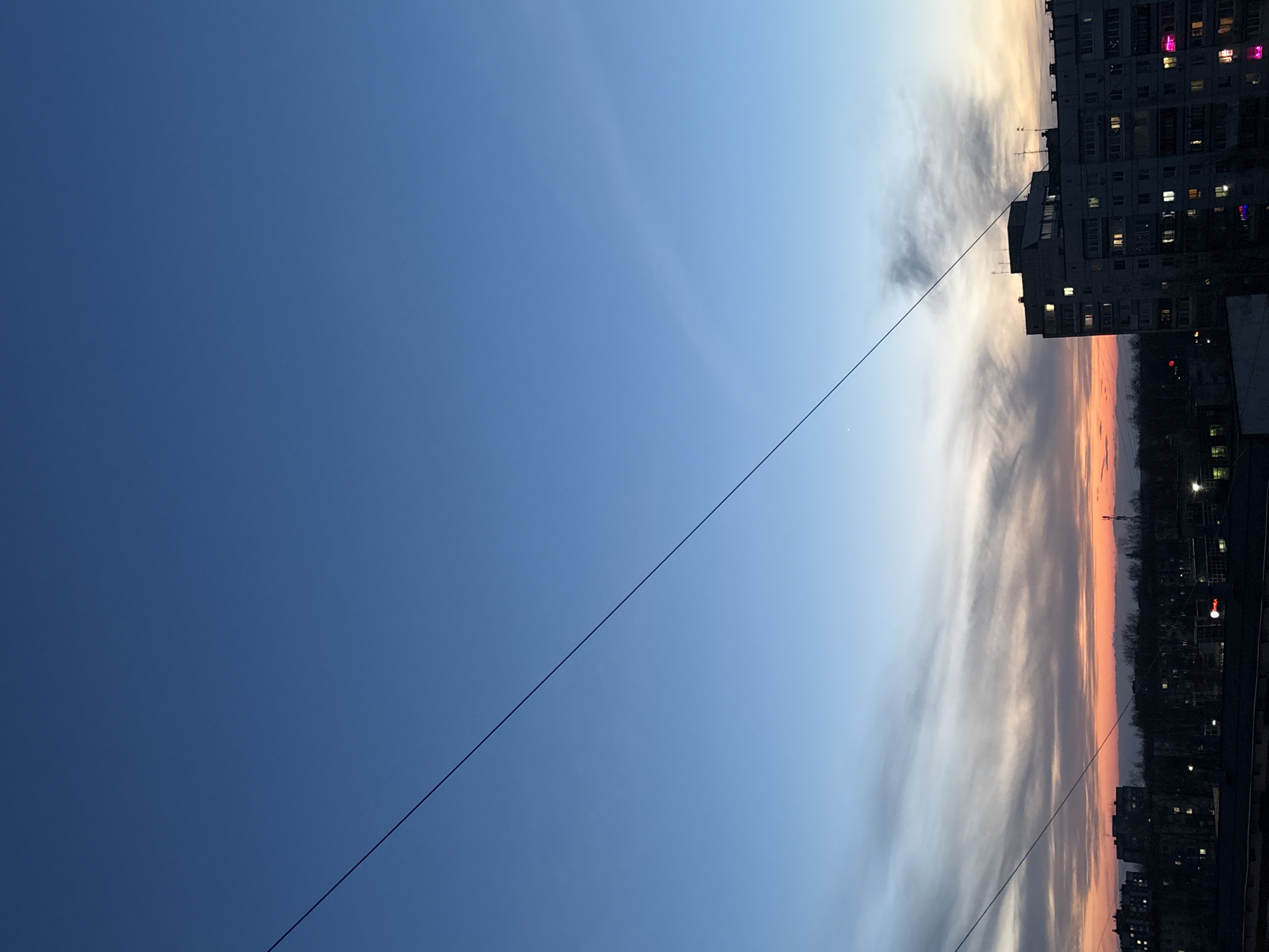 The image depicts a view of the sky during a sunset in the city of Chaikovsky, Russia. The sky transitions from a deep blue on the left to a lighter blue and then to orange and pink hues on the right where the sun is setting. A power line stretches diagonally across the frame. On the right side, silhouetted against the sunset, are multi-story buildings with visible windows, some of which are illuminated. Trees are also visible in the background. The image is oriented vertically.