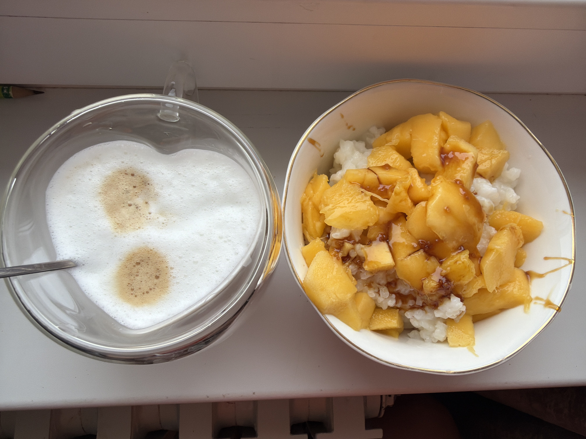 The image shows a top-down view of a glass mug and a bowl placed on what appears to be a white window sill or surface. The mug contains a beverage with a frothy, heart-shaped design on top, possibly a latte or cappuccino, with a spoon visible in the drink. The bowl contains a dessert of what appears to be sticky rice topped with diced mango, drizzled with a sauce. A pencil is visible in the upper left. The photograph was taken in Chaikovsky, Russia.