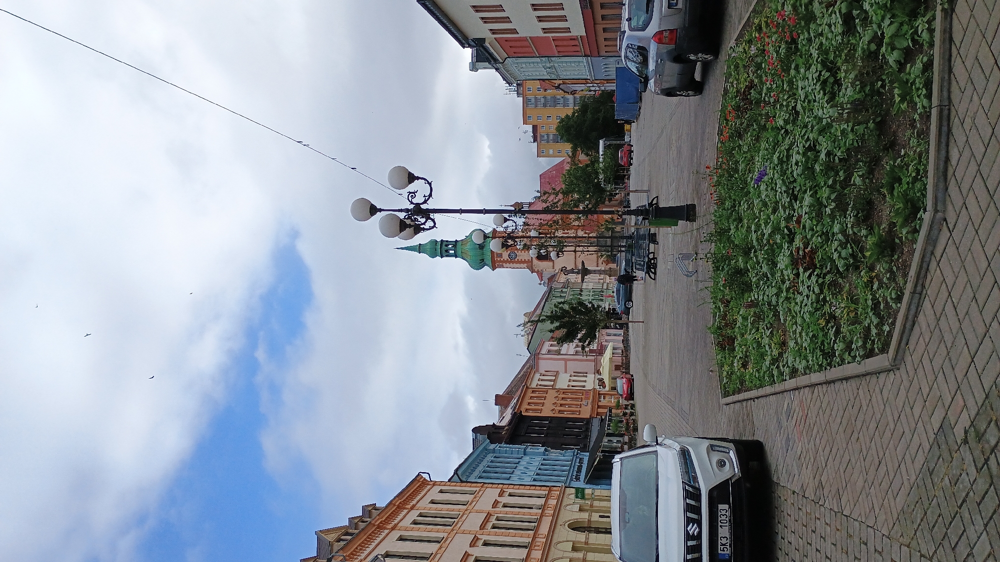 The image captures a view of the Sokolov city square in the Czech Republic. In the foreground, a white SUV is parked on a brick-paved area. Adjacent to the parking area is a well-maintained flower bed filled with green foliage and scattered red and purple flowers.
Dominating the midground is a large, ornate street lamp with multiple globes, casting light over the square. Beyond the lamp, a cobblestone plaza stretches out, lined with a variety of colorful buildings in shades of orange, blue, black, and green. A prominent church with a green, pointed spire stands tall in the center of the square, adding a historical architectural element. Several trees are planted along the walkway, providing some shade.
In the background, more residential buildings are visible, including a taller, modern apartment complex with orange and yellow facades. People can be seen walking in the distance, and other cars are parked or driving through the square. The sky above is partly cloudy with patches of blue sky. Several birds are visible in flight against the sky. The overall impression is of a bustling town square with a mix of historical and modern architecture.