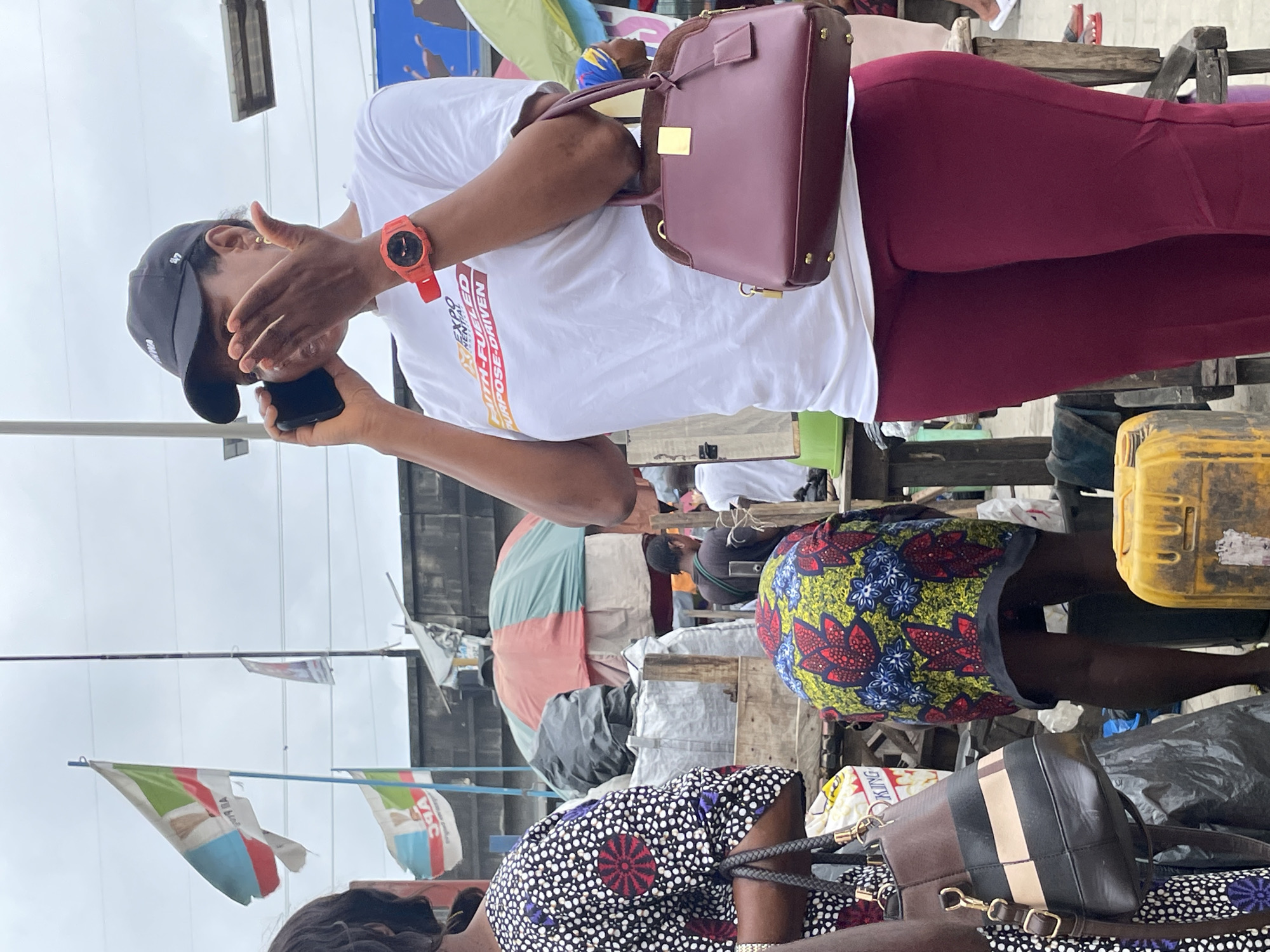 The image depicts a street scene, likely in Lekki, Nigeria. A woman in the foreground is wearing a white t-shirt with text that reads "FAITH-FUELED PURPOSE-DRIVEN" and dark red pants. She has a black baseball cap on and is holding a cell phone to her ear with her right hand, while her left hand is covering her mouth. She is also wearing a red digital watch on her left wrist and carrying a burgundy handbag. To her left, another woman is visible from the shoulders up, wearing a patterned dress. In the background, several flags are flying, including one with the colors light blue, white, green, and red, and the letters "APC". There are also market stalls with umbrellas, bags, and other goods. People are milling about in the background. A yellow plastic container is visible in the lower right corner. The sky is overcast.