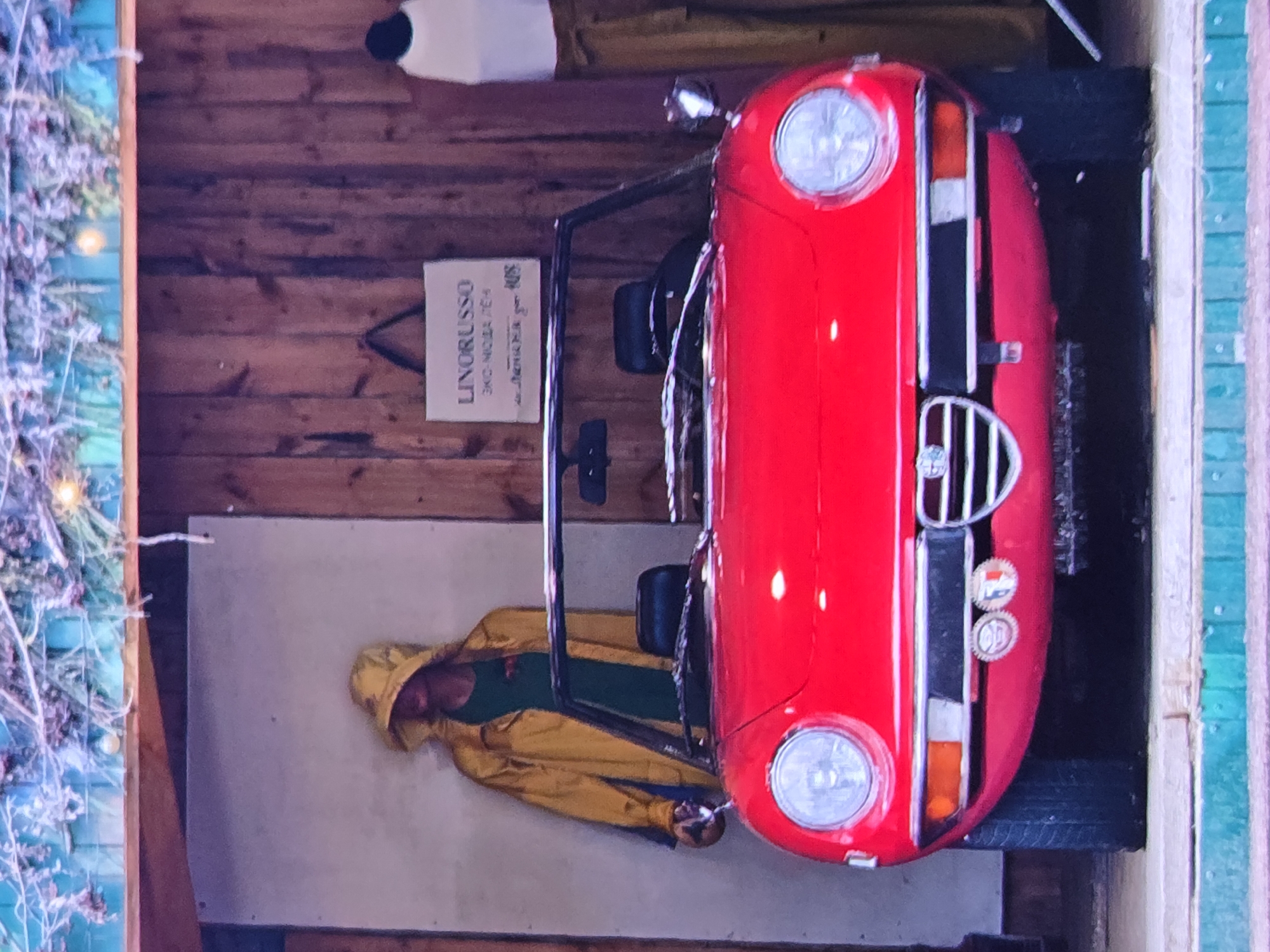 The image shows a red convertible sports car parked in front of a display. To the left of the car, a mannequin wearing a yellow hooded raincoat over a green shirt stands against a light-colored wall. Behind the car, a wooden wall is visible with a hanging sign that reads "LINORUSSO EKO-MODA 7em". To the right of the car, another mannequin is partially visible, wearing a white top and brown pants. The background suggests an indoor or covered outdoor setting, possibly a shop or exhibition space, with overhead decorations. The foreground shows a curb with turquoise detailing. The overall impression is that of a retail display or a decorative arrangement. The image does not contain any specific visual cues to place it in the city of Vladimir, Russia.