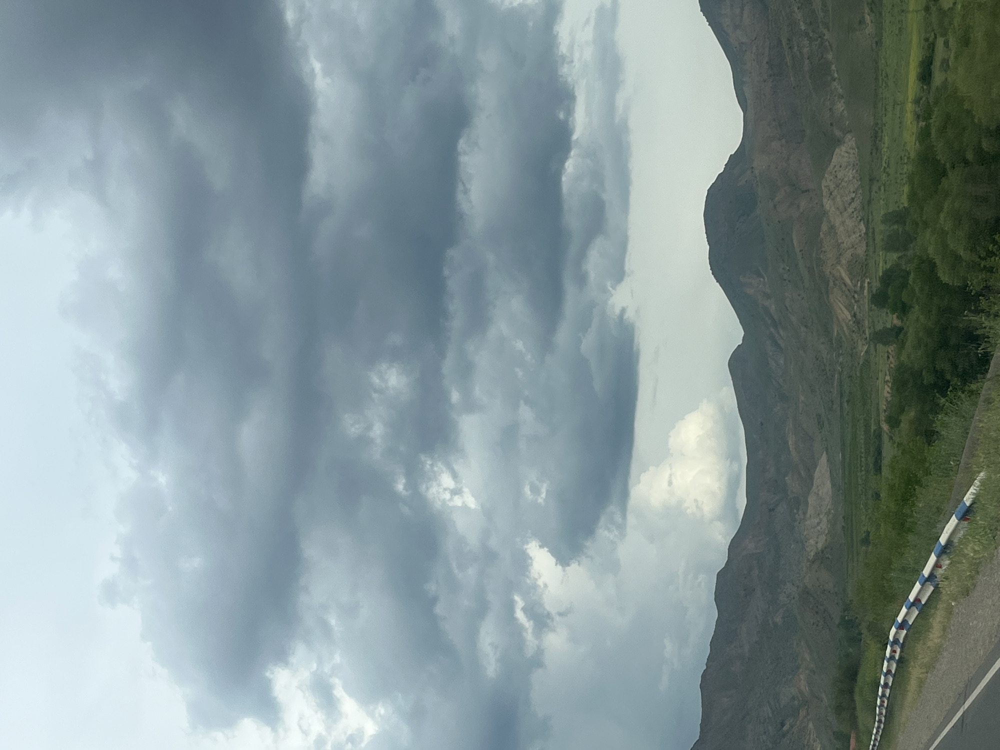 This image captures a dramatic mountainous landscape in Chiva, Armenia, under a heavily clouded sky. The scene is dominated by a rugged mountain range on the right side, characterized by steep, earthy slopes with patches of green vegetation, suggesting a valley or gorge. Further down the mountain and extending into the middle ground, there are flatter areas of vibrant green, possibly agricultural fields or natural grasslands.
The sky is a prominent feature, filled with thick, dark grey clouds that cover most of the frame, hinting at overcast weather or an approaching storm. Patches of lighter sky are visible, particularly on the left, but the overall impression is one of low-hanging, weighty clouds.
In the lower right corner, a segment of a paved road is visible, running alongside the mountain. A distinctive guardrail lines the edge of the road, featuring a pattern of blue, white, and red stripes. No people or specific activities are discernible, giving the impression of a serene yet imposing natural environment. The time of day appears to be daytime, though the heavy cloud cover diffuses the light. No visible text is present in the image.