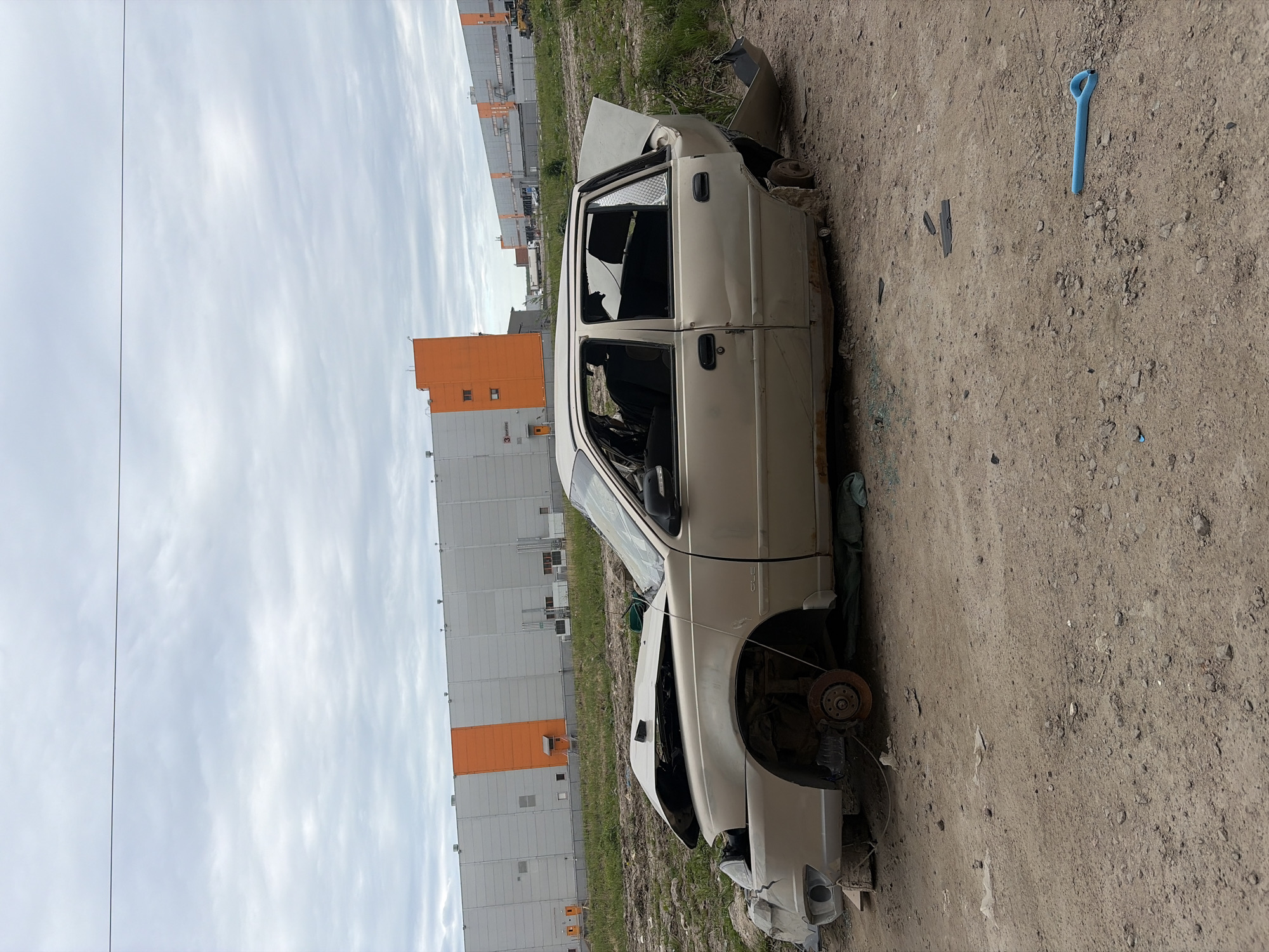 The image shows a heavily damaged white car in an outdoor location. The car appears to be a total loss with broken windows, missing wheels, and significant body damage. The background features a large warehouse or industrial building with grey and orange panels. To the right of the car is a blue plastic object on the dirt ground. Based on the architecture and background details, the location is plausibly in Saint Petersburg, Russia.