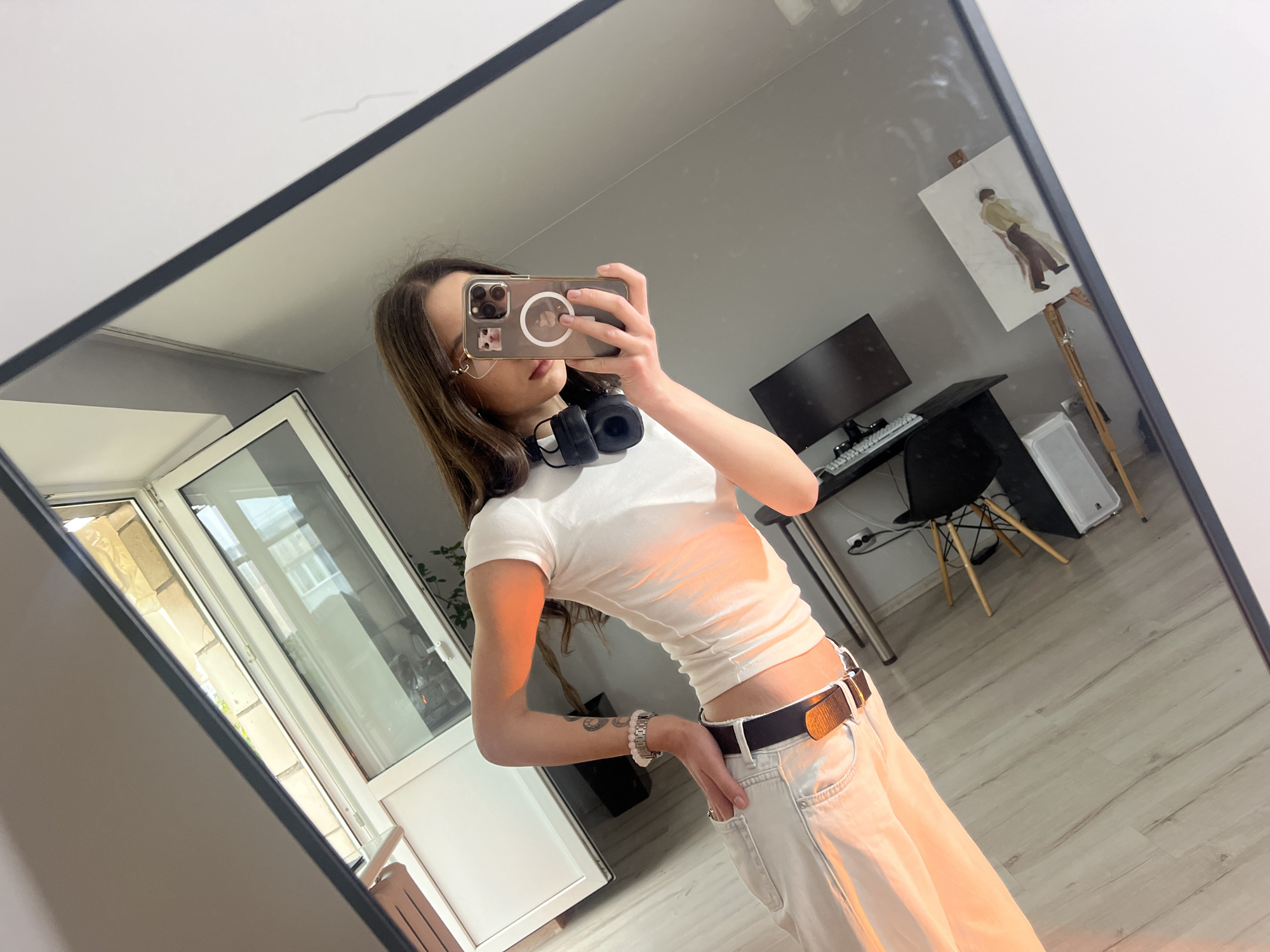 A mirror selfie captures a young woman posing indoors. She is reflected in a large, dark-framed mirror, holding a smartphone with a clear case to take the picture. Her face is partially obscured by the phone, but glasses, earrings, and long dark brown hair are visible. She wears a white short-sleeved crop top, light-colored wide-leg pants, and a dark belt. Black over-ear headphones are draped around her neck. On her left wrist, she has a watch and two bracelets, and a tattoo is visible on her forearm. Her left hand is casually placed in her pant pocket.
The background, also reflected in the mirror, shows a contemporary room with light gray walls and light-colored wooden flooring. To the left, a white-framed glass door or window looks out onto an exterior area with visible brickwork. To the right, a workstation is set up with a dark desk, a black monitor, and a white keyboard. A black chair with wooden legs is pulled up to the desk. Further right, an easel holds a canvas with an unfinished painting depicting a figure in brown pants and a beige top. A white cabinet or shelf unit is visible near the window, and another white rectangular object rests on the floor.
The lighting is bright, indicating daytime. The scene suggests a personal space, likely a bedroom or living area, within a residential setting in Ternopil, Ukraine. No specific text is clearly legible in the image.