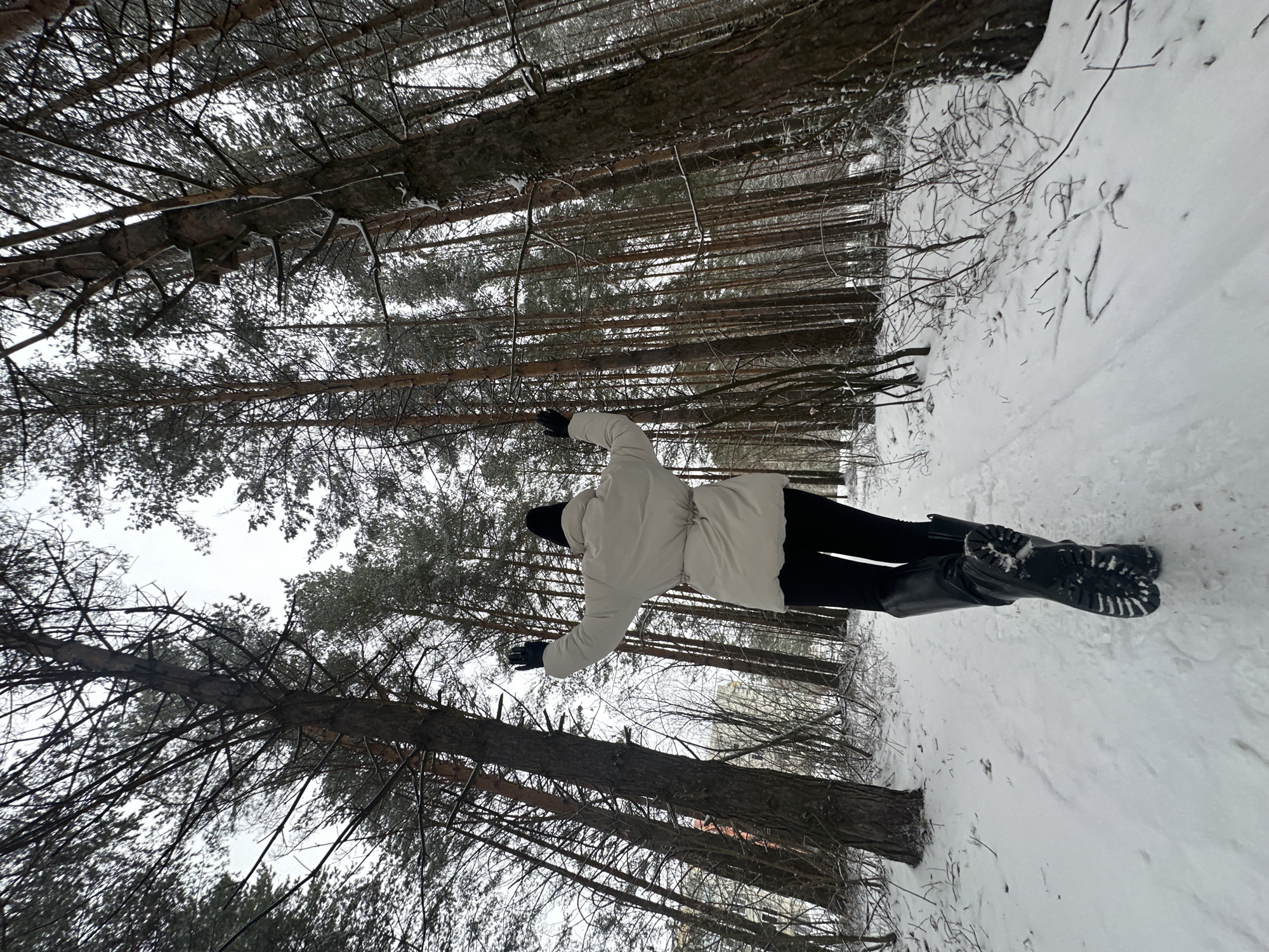 A person is standing in a snow-covered forest in Chaikovsky, Russia. The person is wearing a white jacket, black pants, and black boots. The person has their arms outstretched, as if they are embracing the forest. The trees are tall and bare, and the ground is covered in a thick layer of snow. The sky is overcast, and the light is dim.