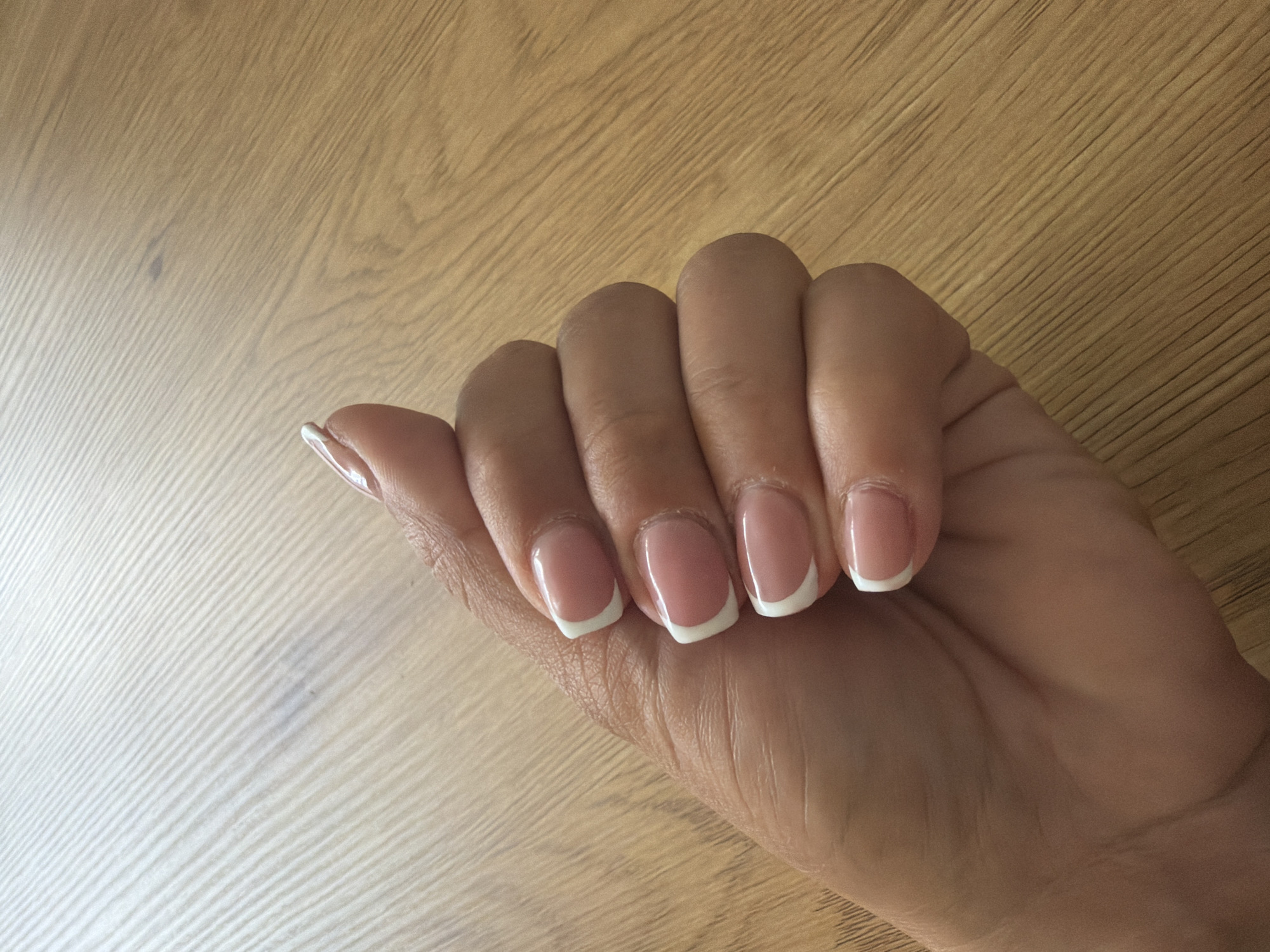 A person's right hand with a French manicure is shown resting on a light-colored wooden surface. The hand is positioned with fingers slightly curled, showcasing the neatly done nails. The nails are of medium length, with a squarish or squoval shape, featuring opaque white tips and a translucent pink or nude base. The skin tone of the hand is a medium shade. The wooden surface, possibly a table or floor, has a visible grain pattern running diagonally across the image, from the top left to the bottom right. There are no other objects or people visible, and no text is present. The lighting suggests an indoor setting. This image is from Svetovrachene, Bulgaria.