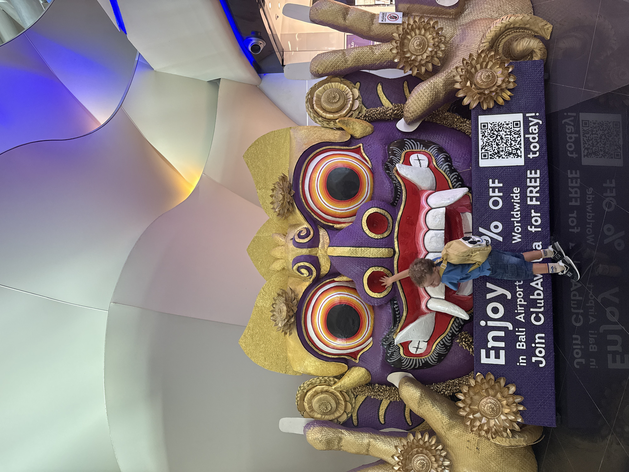 The image depicts a young boy, wearing a blue t-shirt, denim shorts, and a yellow backpack, reaching out to touch a large, vibrant purple and gold sculpture of a mythical creature's face. The sculpture has prominent red lips, white fangs, and large, multi-colored eyes with black pupils. Golden hands with intricate floral designs frame the face. Below the sculpture, a purple banner displays white text that reads, "Enjoy % OFF in Bali Airport Join ClubAvia Worldwide for FREE today!" A QR code is also visible on the banner. The background features a modern, curved architectural design with soft lighting in shades of white, green, yellow, and blue, suggesting an indoor public space like an airport terminal. The floor is a dark, reflective surface. This scene is located in the city of Kuta, Indonesia, specifically within Bali Airport, as indicated by the text on the banner.
