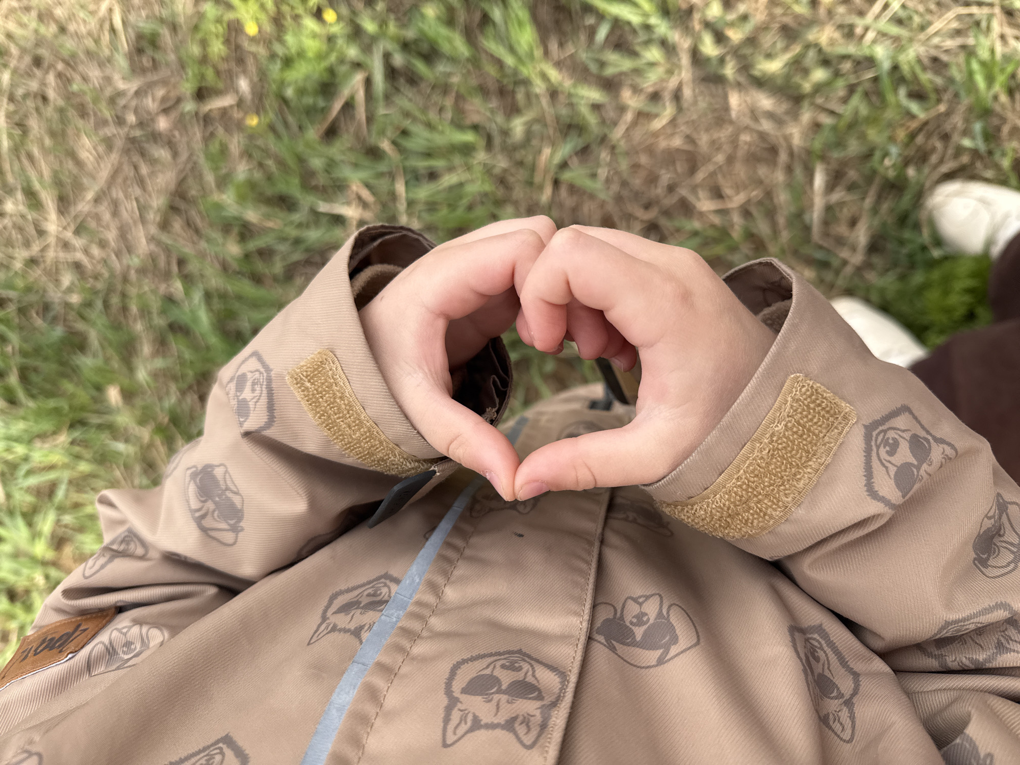 A child stands in a grassy area, possibly in Luzhkovo, Russia. The child is wearing a beige jacket adorned with repeating stylized raccoon or dog-like face patterns. They are holding their hands together to form a heart shape in front of their chest. The background consists of green and brown grass. A glimpse of a white shoe can be seen in the background to the right.