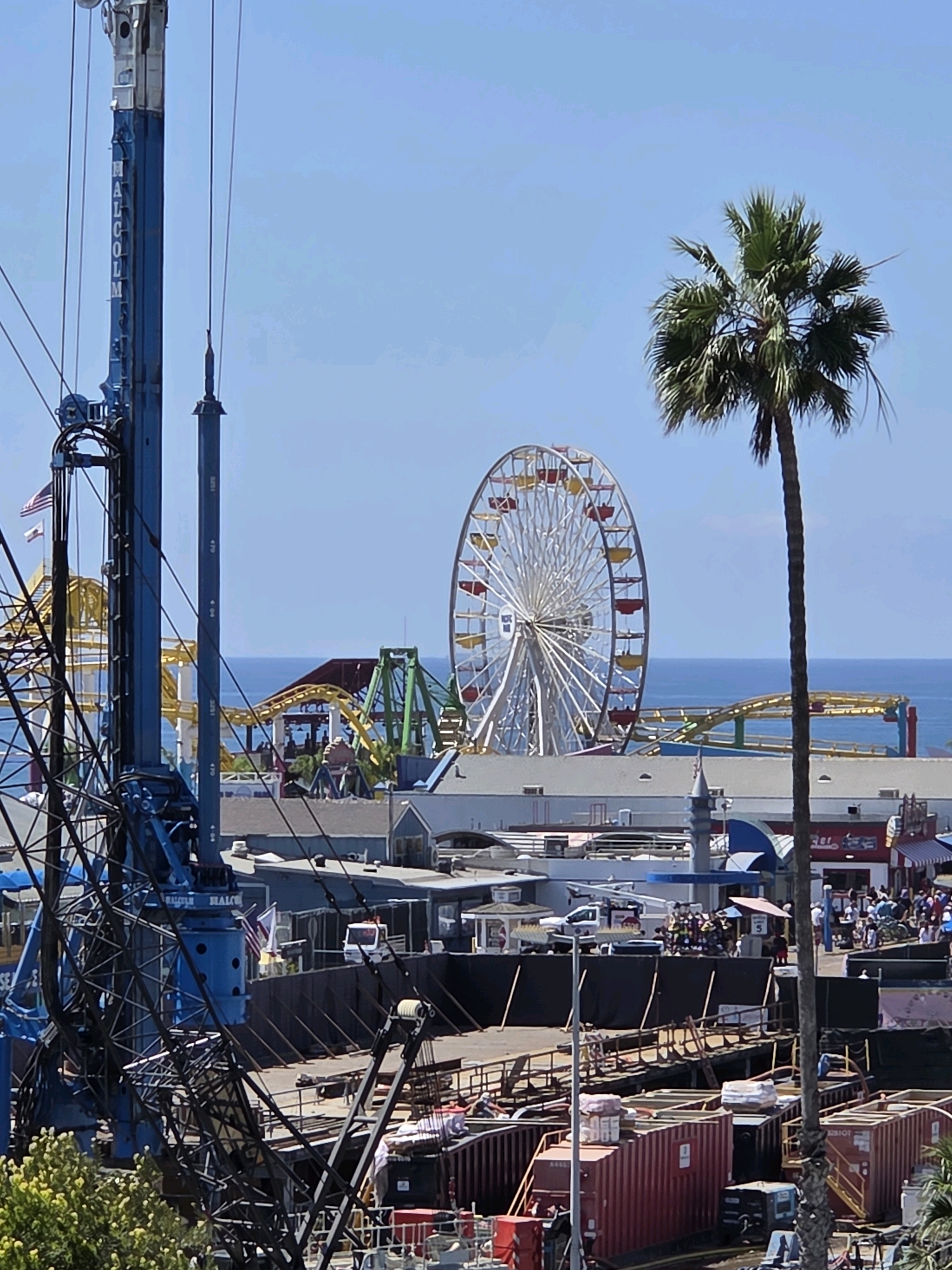 This image captures a daytime scene at the Santa Monica Pier in Santa Monica, California, United States of America. The foreground is dominated by a large blue construction crane, its intricate rigging and cables obscuring a portion of the view. Beyond the crane, the iconic Santa Monica Pier amusement park is visible, featuring the Ferris wheel and the roller coaster. The ocean serves as a backdrop to the pier and the clear blue sky stretches above. The scene appears to be under construction, as evidenced by the presence of shipping containers and barriers in the foreground and midground. A palm tree stands tall to the right of the frame, adding a tropical element to the coastal setting. In the midground, a crowd of people can be seen enjoying the amusement park attractions. The overall atmosphere is one of a vibrant, sunny day at a popular tourist destination, with a hint of ongoing development.