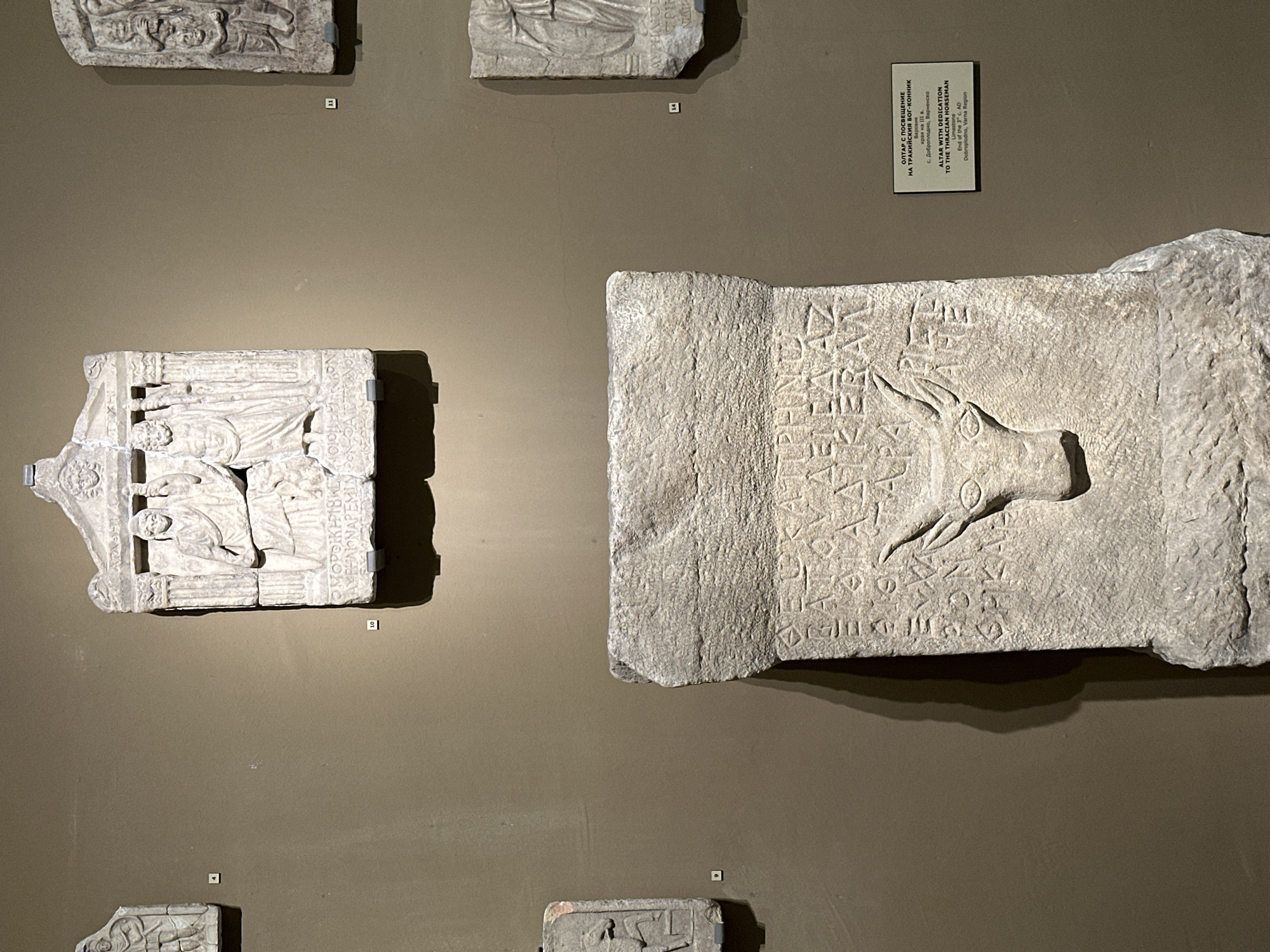 This is a medium-shot, eye-level photograph of an archeological display in the city of Varna, Bulgaria. The background is a plain, textured wall in a muted brown color. In the foreground, a large, rough, stone altar is positioned centrally. The altar is rectangular and appears to be broken at the top and bottom. The front face of the altar is carved with ancient inscriptions and a relief carving of a bull's head with horns. The bull's head faces forward and is positioned in the lower half of the altar. Above the bull's head, rows of ancient text are carved into the stone, some of which is illegible. To the right of the altar, a small, rectangular information plaque is attached to the wall. The plaque is white with black text and is bilingual, containing information in both Bulgarian and English. The English text reads: "ALTAR WITH DEDICATION TO THE THRACIAN HORSEMAN, Varna Region, End of the 3rd c. AD, Dobroplodna, Varna Region".
On the wall above and to the left of the altar, there are several smaller stone artifacts displayed. One artifact, labeled with the number 4, is a rectangular stone slab with a relief carving of a standing figure in a toga. To its right, another, larger rectangular stone artifact labeled with the number 10, features a pediment-like top with faces carved into it and a relief of two figures standing within columns. Below these figures, an inscription is carved into the stone. To the right of artifact 10, another artifact labeled with the number 11, shows a relief of several figures stacked vertically. Further to the right and slightly above artifact 11, a portion of another stone artifact is visible. To the right and below artifact 11, a rectangular stone artifact labeled with the number 14, exhibits a relief carving of a standing figure. The lighting in the display case is somewhat dim, with spotlights illuminating the artifacts. The overall impression is of a museum or gallery setting showcasing ancient historical objects.