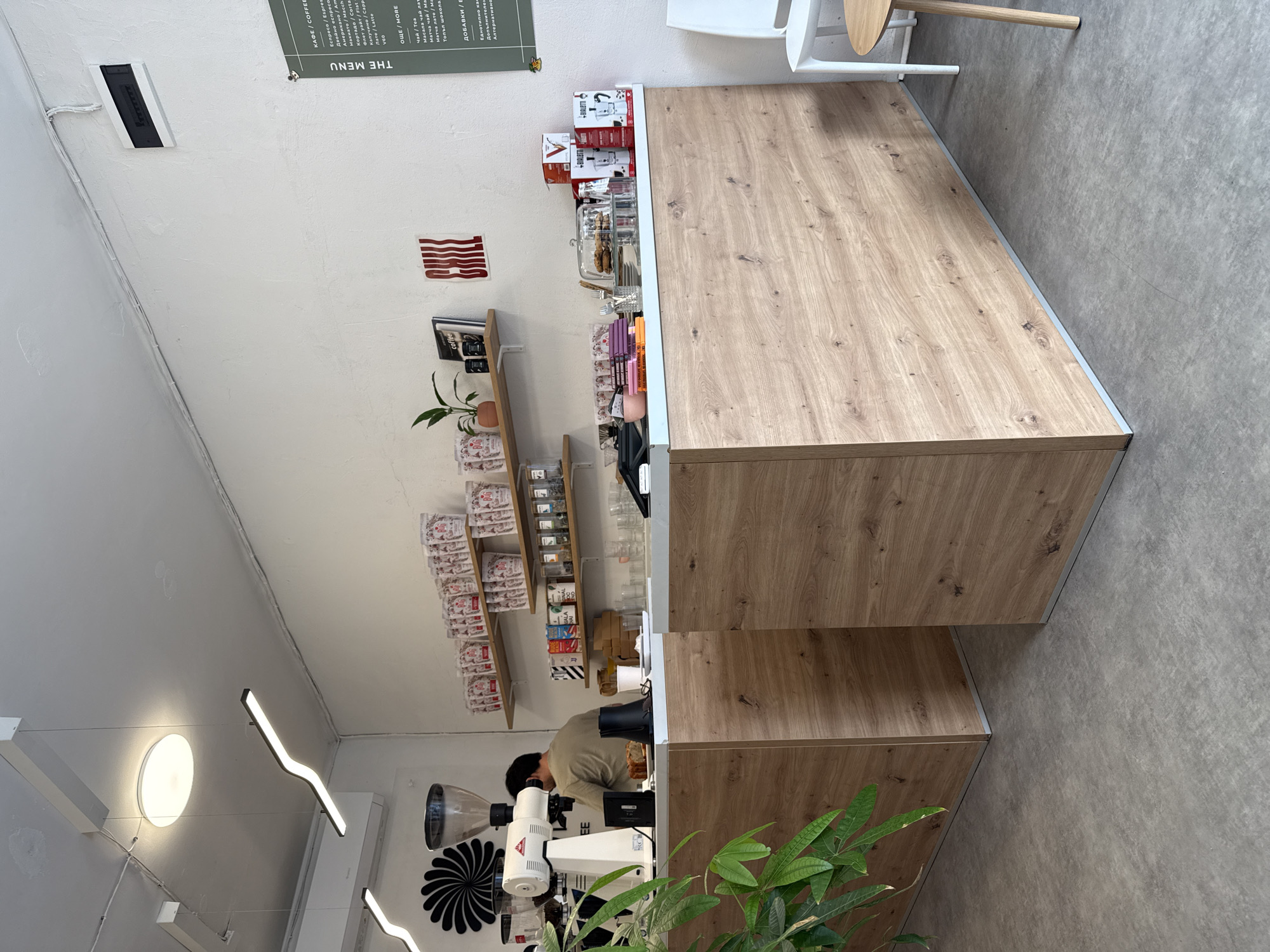 This image captures the interior of a modern coffee shop or cafe in Sofia, Bulgaria, characterized by its minimalist and industrial-chic aesthetic. The central focus is a large, light-wood counter with a subtle grain, serving as the main service area. Behind the counter, a barista is visible, dressed in a light-colored shirt, operating a white coffee machine and grinder, suggesting they are in the process of preparing a beverage.
On the counter, various items are arranged, including small pastries under a glass dome, colorful notebooks, and boxes of BIALETTI coffee makers, indicating products for sale. The walls are painted white, providing a clean backdrop for the wooden shelves displaying an assortment of packaged goods, coffee beans, and small plants in terracotta pots. A prominent green menu board, titled "THE MENU" in both English and Bulgarian (КАФЕ / COFFEE), lists various coffee drinks like Espresso, Americano, Cappuccino, Latte, and other items like Tea, Matcha, and Hot Chocolate. An additional section for "ДОБАВКИ / EXTRAS" is also present.
Notable decor includes a stylized red wall art piece, resembling abstract strips or text, and a book titled "coffee" on a shelf. The lighting fixtures are contemporary, with sleek linear LED lights and a large circular flush-mount light on the ceiling, illuminating the space brightly. The floor is a neutral grey, resembling concrete, complementing the industrial design. A vibrant green plant in the foreground adds a touch of nature. The overall impression is a bright, functional, and inviting environment for coffee enthusiasts.