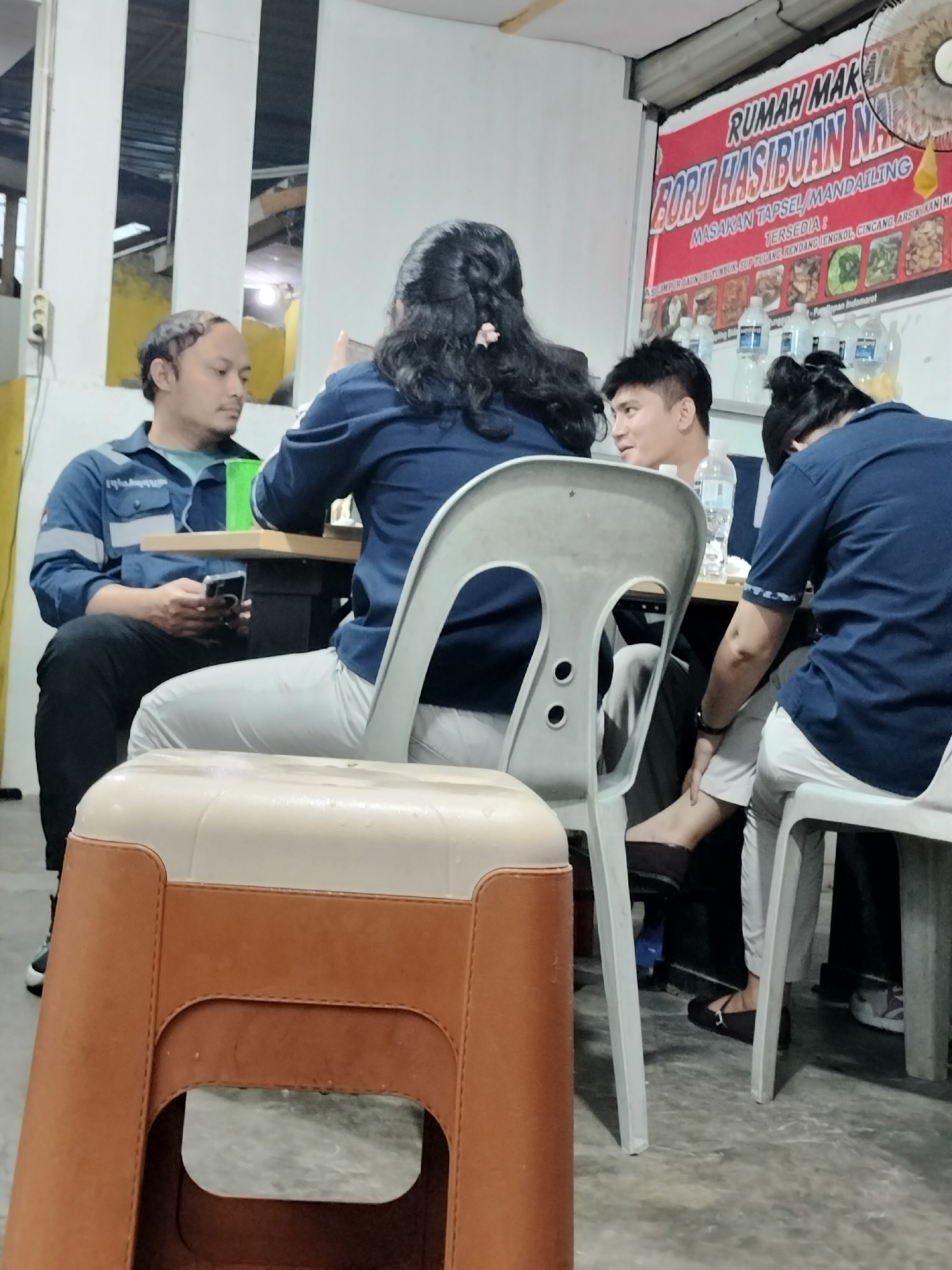 In Batam, Indonesia, a group of individuals are seated at a table in a casual dining establishment. A man in a blue work shirt holds a smartphone. A woman with long dark hair is seated opposite him. To their right, another man is looking away from the camera. A fourth person, also in a blue shirt, is partially visible. Behind the seated individuals, a red banner advertises "RUMAH MAKAN BORU HASIBUAN" and lists various food items. Several bottles of water are placed on the table. The environment appears to be an indoor eating area with a concrete floor and industrial-style lighting. A beige and brown plastic stool is positioned in the foreground.