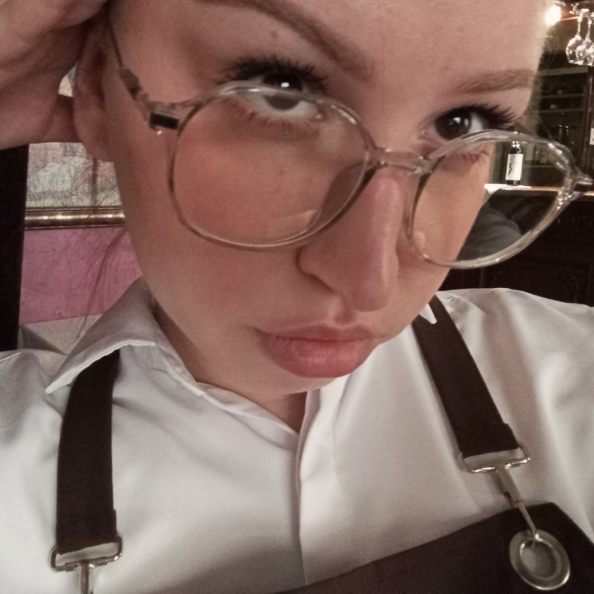 A close-up selfie shows a young woman with light skin wearing round eyeglasses with transparent frames. She has visible eyebrows, eyelashes, and a slight frown. Her hair appears blonde and pulled back.
She is dressed in a white collared shirt with dark brown apron straps over her shoulders, held in place by silver metal attachments.
The background is slightly blurred, but elements suggest an indoor setting. A framed picture is visible to her left. A wine bottle and glasses can be seen behind her to the right.
There is no information to determine the location is in Chaikovsky, Russia, and there is no information to suggest that this is the location.
