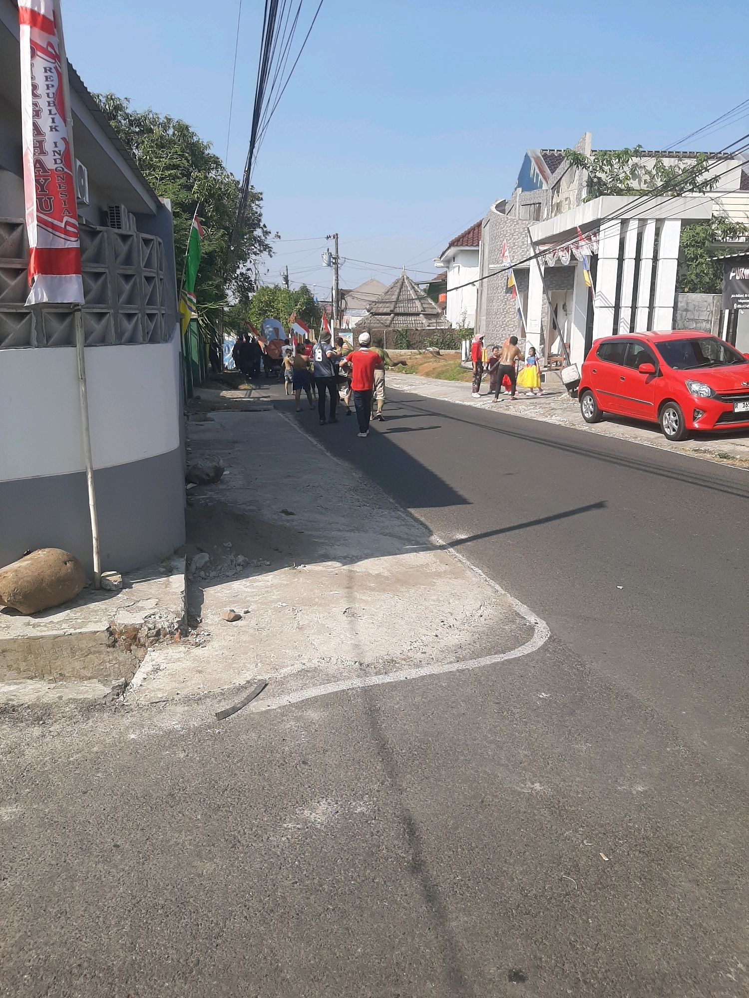 The image captures a street scene in Sumampir, Indonesia, under a clear blue sky. In the foreground, a cracked concrete sidewalk runs along the left side of the asphalt road. A large, round stone rests on the sidewalk near a damaged section of the concrete. A red car is parked on the right side of the road. A group of people, some in red shirts, are walking down the street in the middle ground, appearing to be part of a procession or gathering. Several Indonesian flags are visible, attached to poles and banners on buildings and held by some of the individuals. The architecture includes modern buildings with white columns and patterned facades. The environment suggests a residential or mixed-use urban area.