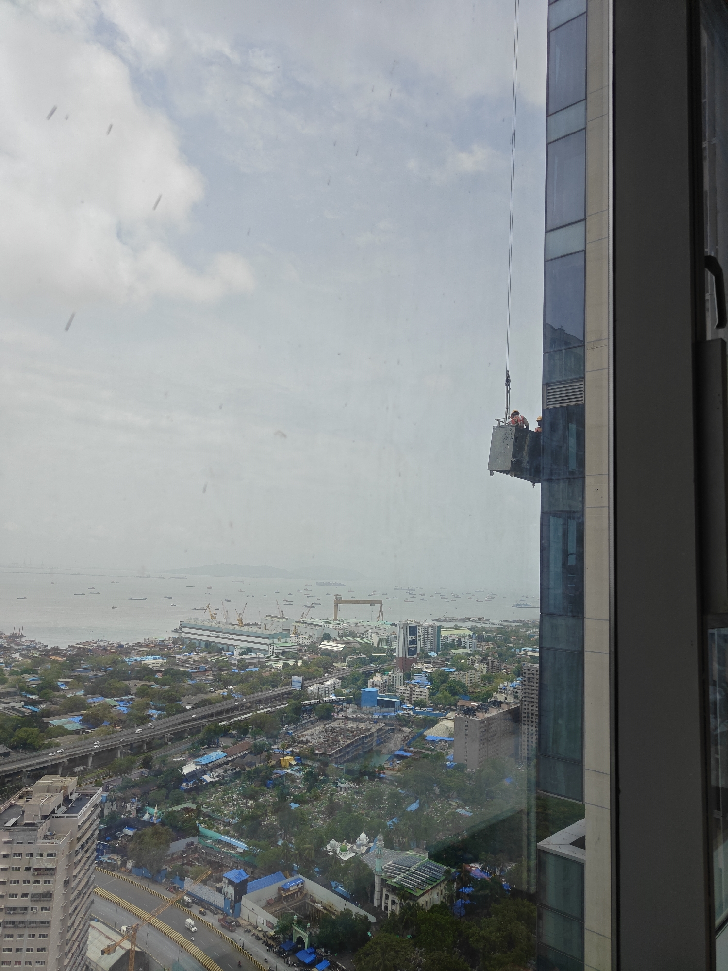 An elevated view from a building in Mumbai, India, shows a large body of water with numerous ships in the distance. In the mid-ground, a bustling cityscape unfolds with various buildings, a highway overpass, and industrial structures, including a large shipyard with cranes. Closer to the foreground, a construction site is visible with unfinished buildings and temporary structures. On the right side of the frame, a modern skyscraper dominates, with a suspended platform carrying two workers in orange hard hats, appearing to be engaged in maintenance or window cleaning on the building's exterior. The sky is overcast with clouds.