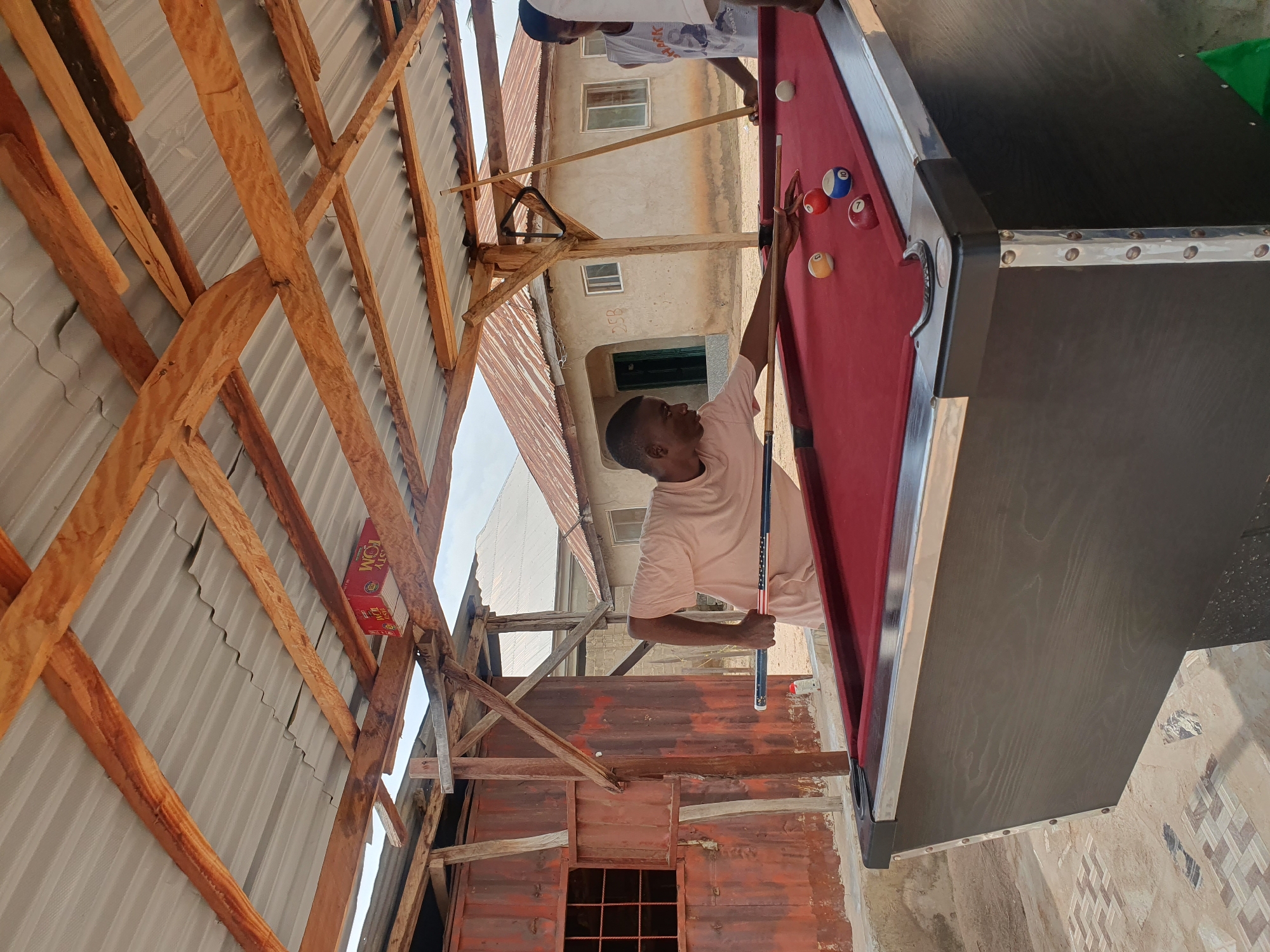 In Lokoja, Nigeria, two young men are playing billiards under a corrugated metal roof supported by rough-hewn wooden beams. The scene appears to be an outdoor recreational area with a red billiard table in the foreground. One man, wearing a light-colored t-shirt, is in the process of taking a shot with a pool cue, aiming at a yellow ball. He is positioned on the left side of the table. Several other billiard balls are scattered on the table, including a red ball with the number '1' and a blue ball with the number '10'. A rack for billiard balls is visible hanging from one of the wooden beams above. In the background, there are rustic structures, including a corrugated iron wall with a barred window on the left, and simple buildings with corrugated roofs and exposed brickwork. Another young man, wearing a white tank top with a graphic print, stands on the right side of the billiard table, holding a pool cue. The ground is paved with decorative tiles in some areas. The overall environment suggests an informal setting.
