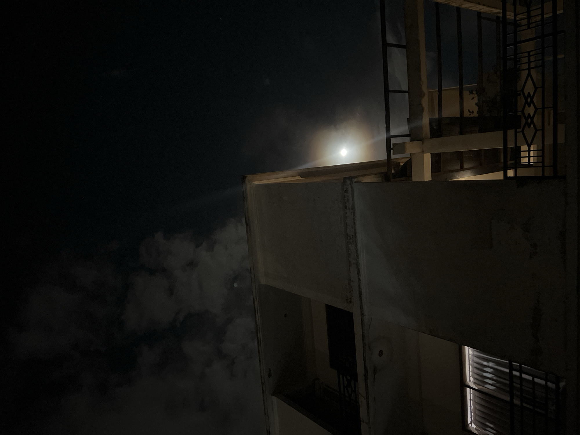 The photograph depicts a nighttime scene, likely in an urban area. The view is directed upwards, showcasing the corner of a multi-story building. A bright moon, partially obscured by clouds, illuminates the top edge of the building. The building's architecture features balconies and what appear to be window slats. The sky is dark, with cloudy areas. No people are visible. It is not possible to determine the location as Canggu, Indonesia based on visual details alone.