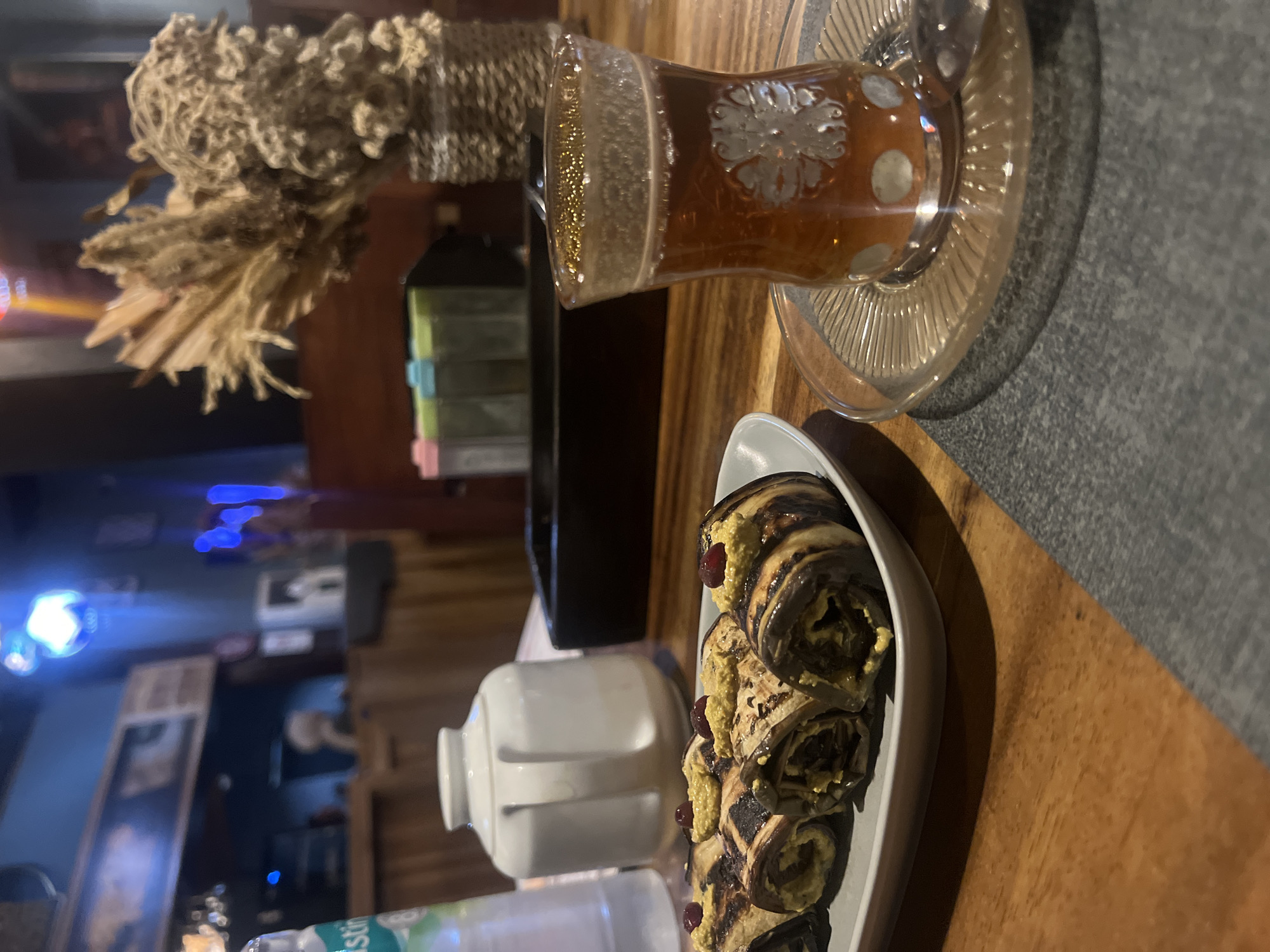 The image shows a table setting in what appears to be a cafe or restaurant. On the table is a glass of tea on a decorative saucer, a white serving dish containing rolled aubergine or eggplant, a small white creamer or teapot, and a water bottle. In the background, there is a decorative dried plant arrangement and indistinct elements of the cafe's interior, including lighting and other patrons. The location is specified as North Kuta, Indonesia, though this is not directly evident from the image itself. No people are clearly visible, and interactions are not observed.