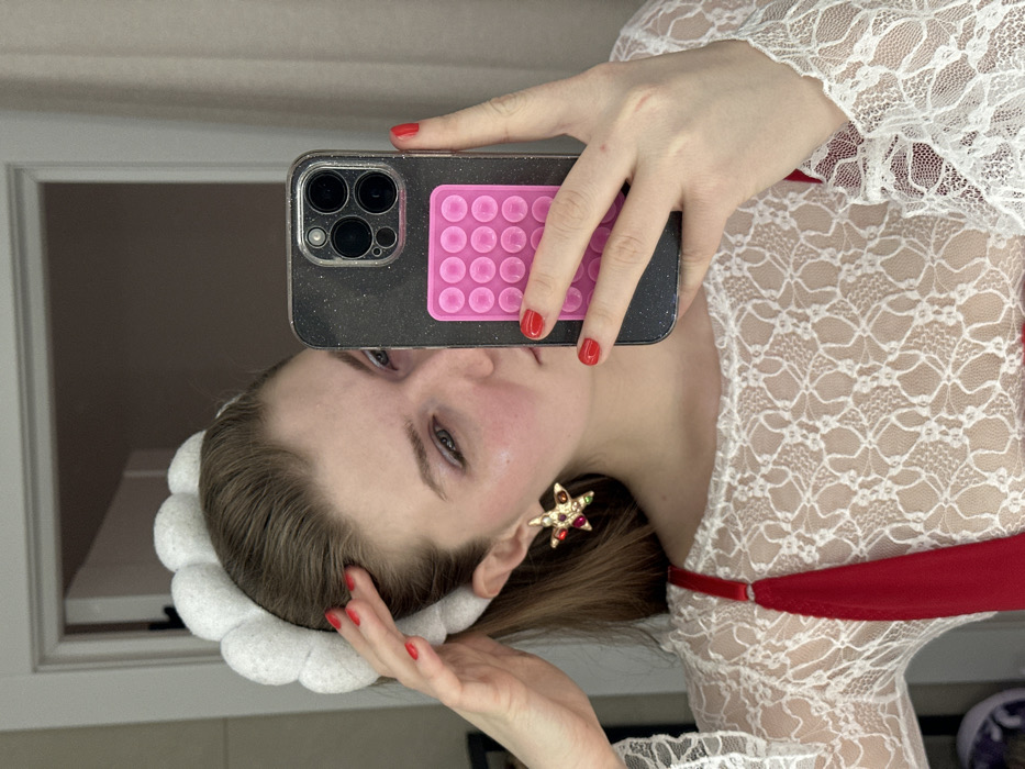 A woman holds a smartphone with a pink "pop-it" on the back, obscuring her face slightly. She wears a white lace top with a red undergarment, and a white fluffy headband. A large, star-shaped earring is visible on her left ear. The woman has fair skin and red nail polish. The background includes a window and a hint of a mirror selfie setting.