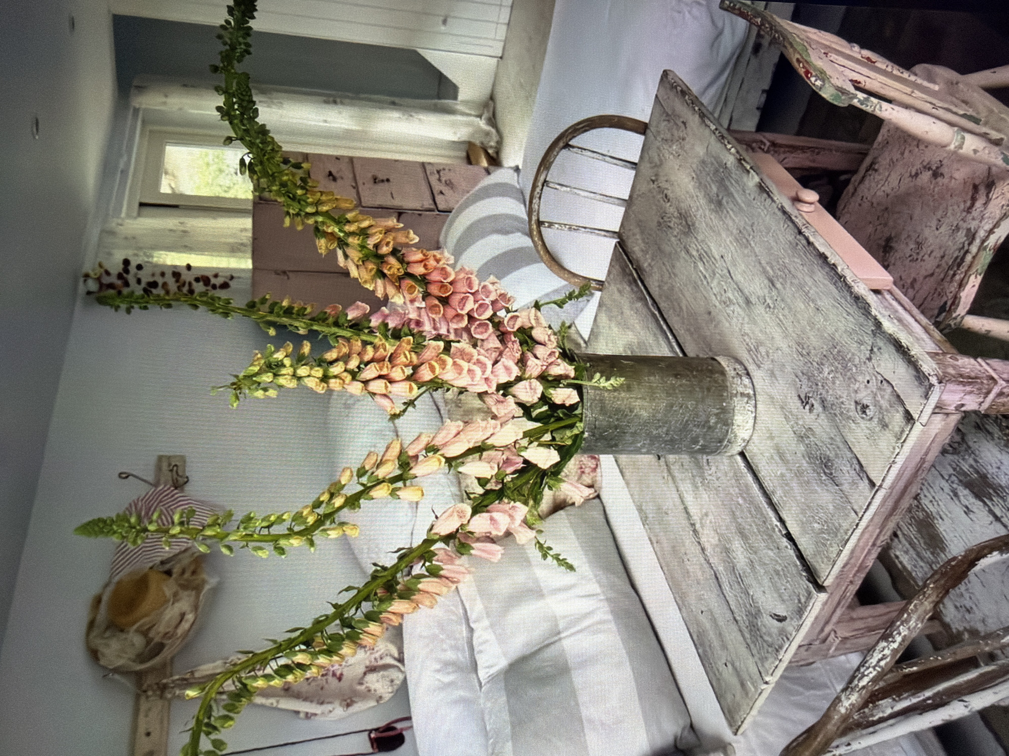 This image captures a serene indoor scene with a rustic, shabby chic aesthetic, likely set in Ros Na Rí, Ireland. The photograph, presented rotated from its original orientation, centers on a bountiful bouquet of pink and peach foxgloves arranged in a weathered, cylindrical metal vase.
The vase rests on a distressed wooden table, painted in a chipped light grey or white, revealing hints of natural wood underneath. Around the table are several vintage-style wooden chairs, similarly aged with peeling paint. One chair features a distinctive curved, rusty metal backrest, adding to the antique charm.
In the background, a window with white frames allows abundant natural light to stream into the room, illuminating hints of green foliage outdoors and suggesting it is daytime. The interior walls are painted a light, subtle blue-grey. Adjacent to the table, a bed or cushioned seating area is visible, covered with a light-colored fabric featuring soft vertical stripes. Another patterned fabric is draped nearby.
To the left of the frame, a rustic wooden wall fixture holds decorative elements, including a piece of striped fabric and a small, light-colored woven item, possibly a miniature hat or decorative basket, with another patterned fabric hanging below.
The scene evokes a sense of tranquility and homely comfort. There is no active event taking place, presenting a peaceful still life or interior shot. No visible text is present in the image.