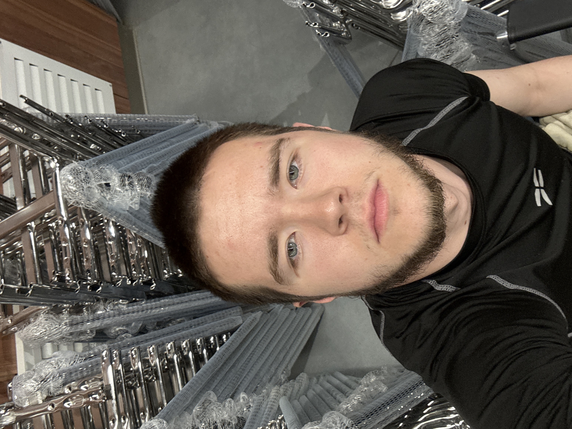 A selfie taken from a high angle of a light-skinned male with brown hair and a beard, wearing a black long-sleeved shirt. The background shows a stack of what appears to be chrome-plated chairs, many still wrapped in protective plastic mesh, suggesting the photograph was taken in a storage area or furniture warehouse. The location in Kazan, Russia, is not discernible from the image.