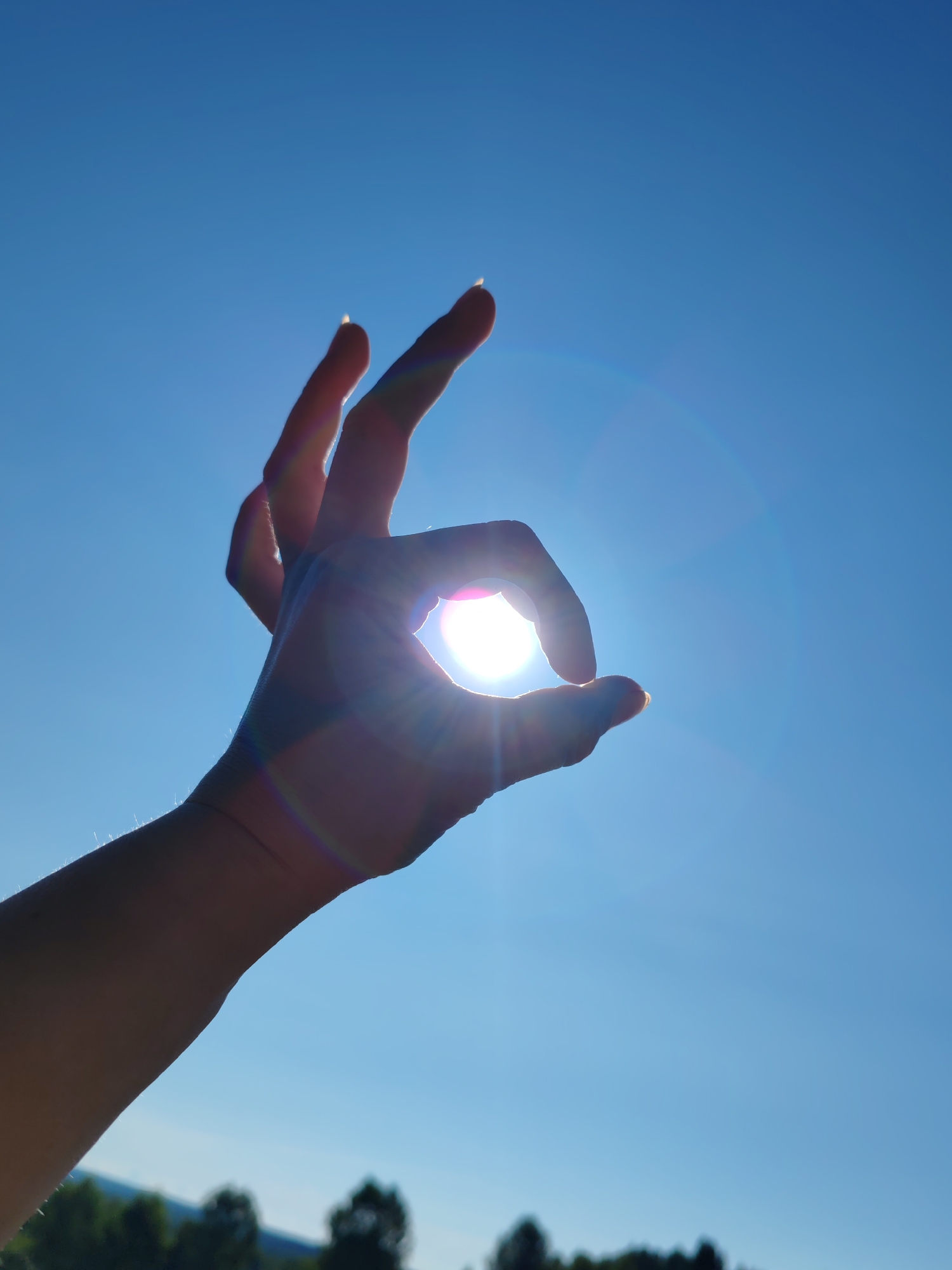 A person's hand is silhouetted against a bright blue sky, forming an "okay" gesture. The sun is partially obscured by the thumb and index finger, creating a lens flare and a rainbow-like effect. The hand is positioned in the foreground, with a slightly blurred background of green trees and hills. The image is taken from a low angle, looking up at the hand and sky.