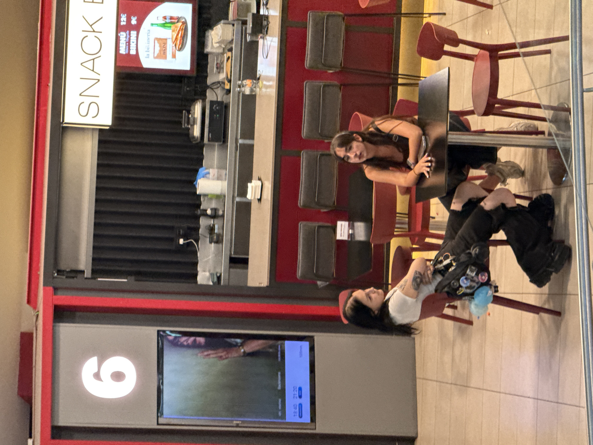 The image depicts an indoor scene that appears to be a snack bar or concession area, likely within a cinema or entertainment venue, given the presence of numbered screens and a "SNACK BAR" sign. The setting is characterized by red and gray color schemes. In the foreground, two young women are seated at separate tables. The woman on the left, with dark hair and tattoos on her arms, is wearing a white t-shirt and black pants, with a bag adorned with various pins hanging from her chair. The woman on the right, also with dark hair, is wearing a dark tank top and jeans. Both are looking away from the camera. In the background, there is a counter with equipment, seating, and posters. One poster displays food items and prices, including a "MENÚ 12€" and "BIKINI 9€". The lighting suggests an indoor environment, possibly with ambient entertainment lighting. The scene is clean and well-maintained. The tiled floor is visible in the foreground. The overall atmosphere is casual and relaxed.