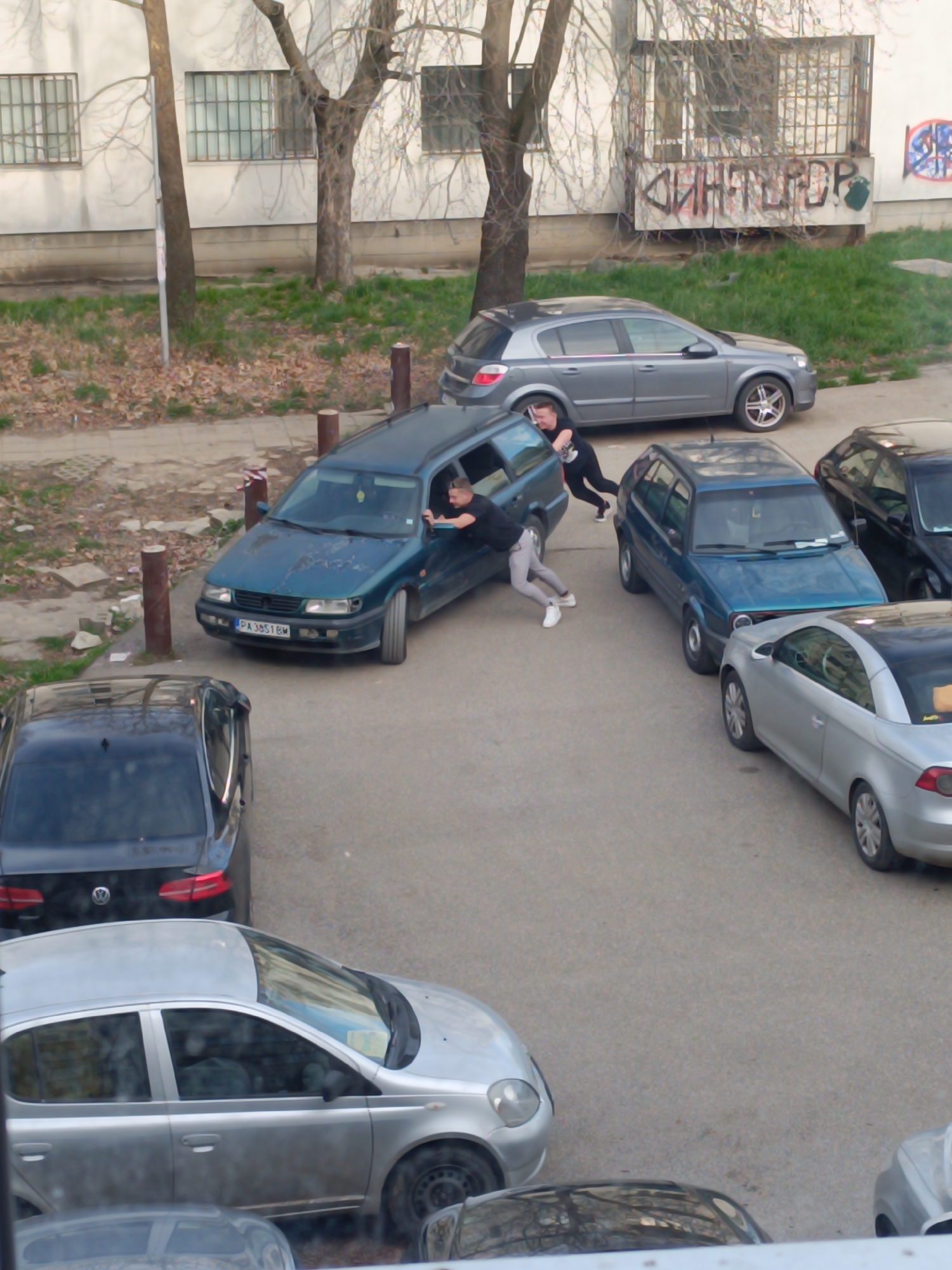 The image shows a parking lot in what appears to be a residential area of Sofia, Bulgaria. The lot is surrounded by apartment buildings and trees. Several cars are parked in the lot, including two men are pushing a blue Volkswagen Golf station wagon. One man is in the driver's seat of the Volkswagen. The license plate of the green car is PA3851BM. Graffiti is present on the balcony of one of the buildings.