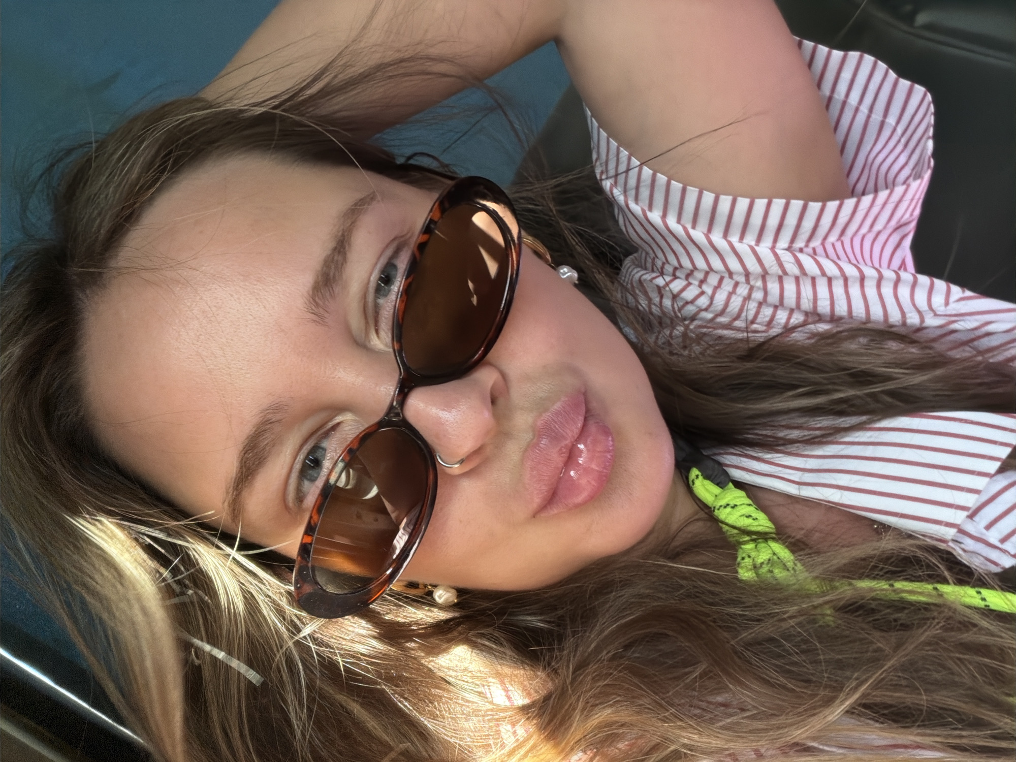 A woman with long brown hair is the central subject of the image. She is wearing sunglasses with brown lenses and a tortoise-shell frame. A silver nose ring is visible in her left nostril, and she has a fair complexion with blue eyes. She is wearing a red and white striped shirt and what appears to be a neon green rope or cord is visible at her neck. The background features a blue surface and dark upholstery, suggesting she is in a car. It is not possible to determine the location of the photo within the city of Dvigatel', Russia.