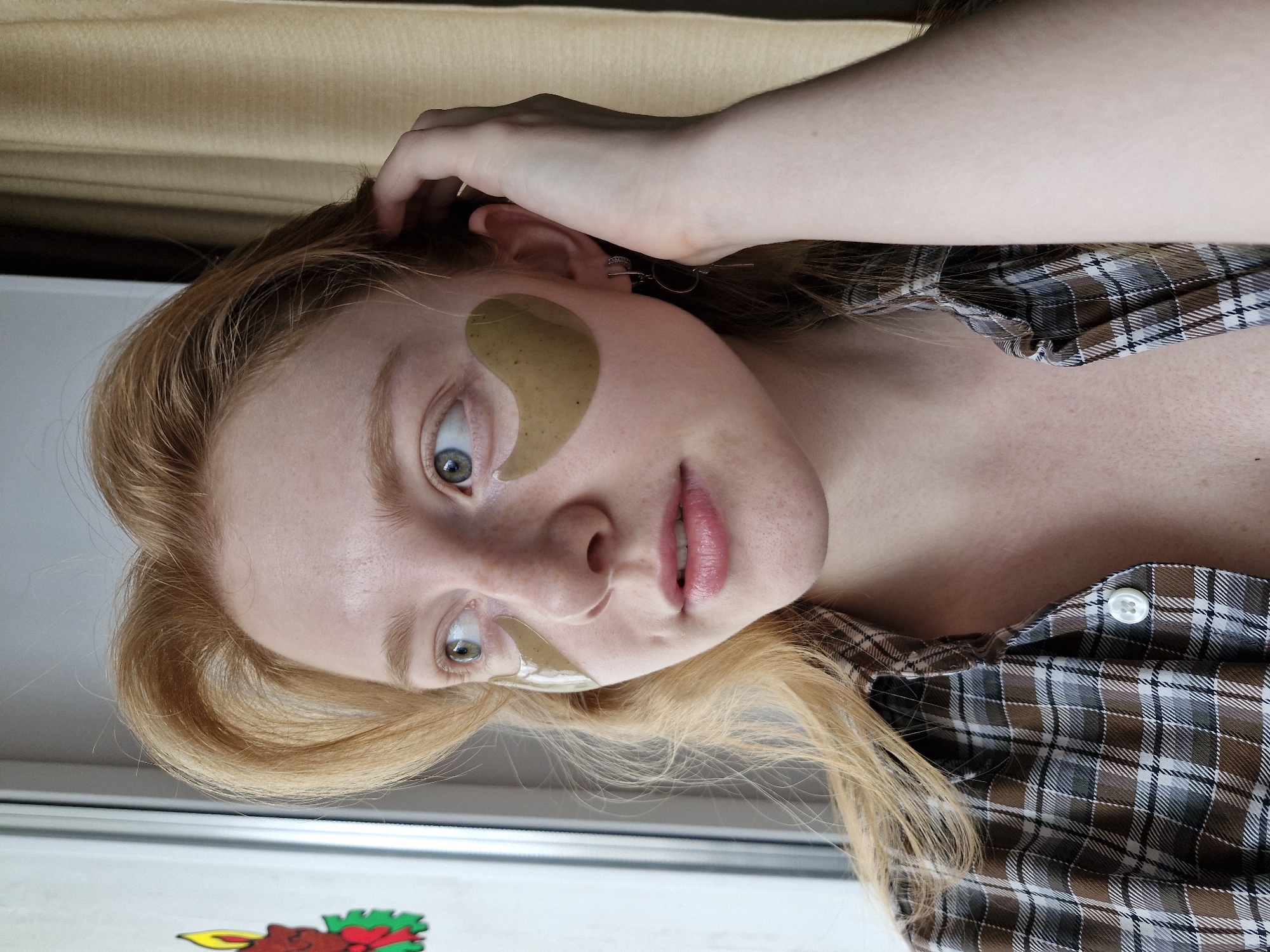 A fair-skinned woman with red hair and freckles is seen from a close, slightly low angle. She is wearing two under-eye skin treatment patches. Her hair is pulled back on one side, and she is wearing a brown and white plaid shirt. The background includes a beige curtain and part of a window frame. A sticker is visible at the bottom left of the frame. The location is not ascertainable from the image.