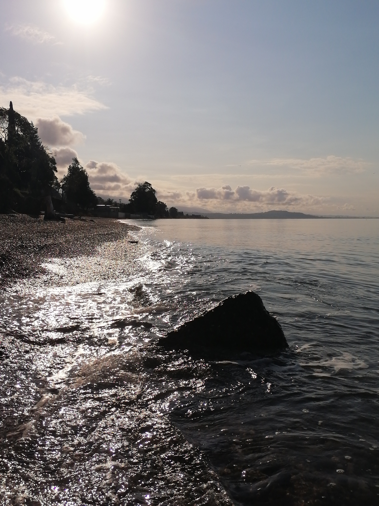 This image depicts a serene coastal or lakeside landscape, likely captured during either sunrise or sunset, evidenced by the bright, low-angle sun glare. The scene is viewed from a pebbly beach, looking out over a calm body of water.
In the immediate foreground, a prominent, dark rock is partially submerged in the water on the right. The water's surface, particularly towards the left near the shore, is covered in countless sparkling reflections from the sun, indicating gentle ripples and small waves lapping at the edge. The water itself appears dark and deep in areas not directly illuminated by the sun.
To the left, a curving shoreline composed of dark pebbles and small rocks extends into the distance. This shore is bordered by a dense line of trees, mostly dark in silhouette, suggesting a verdant bank. Some buildings are faintly visible nestled among the trees further along the shore. Beyond the water, in the far distance, a range of soft, hazy hills or mountains stretches across the horizon.
The sky is a clear, light blue, brightening significantly around the intense sun which is positioned in the upper left of the frame. Scattered, fluffy clouds are visible, with some gathering above the distant landmasses. The overall atmosphere is peaceful and natural. There are no people visible, nor is any text present in the image.