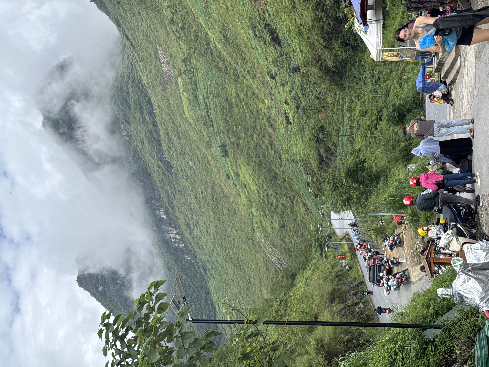 The scene depicts a mountainside in the Yên Minh Commune, Vietnam, with a winding road filled with parked motorbikes and people. The mountainside is lush and green, with some areas shrouded in mist. A woman is standing near a makeshift shop or stall. Further down the road, many motorbikes are parked on both sides. People are gathered around the vehicles, some standing, some sitting. There are small shops or stalls lining the road as well. The sky is overcast, adding to the atmospheric perspective of the landscape.
