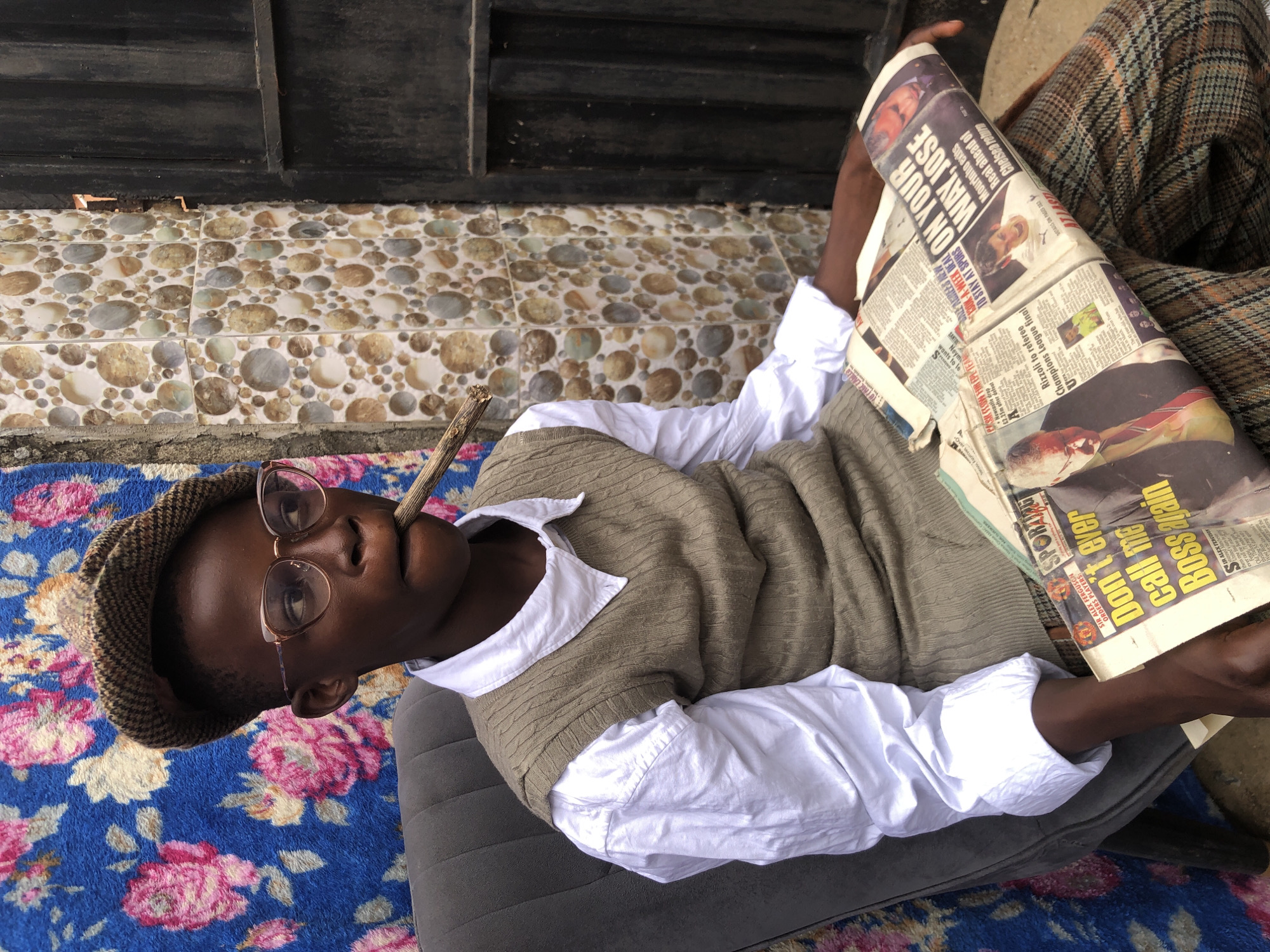A young Black boy wearing a tweed cap, glasses, a white collared shirt, and a brown knitted vest is seated on a grey armchair, holding a newspaper and a prop cigar in his mouth. He is looking to his right. Behind him is a blue tapestry with pink floral patterns, and to his right is a wall decorated with circular patterned tiles and a black metal gate. The boy is dressed in a newsboy style, with plaid trousers visible. The newspaper he is holding displays headlines related to football. The image appears to be taken outdoors. The scene is set in Ado, Nigeria.
