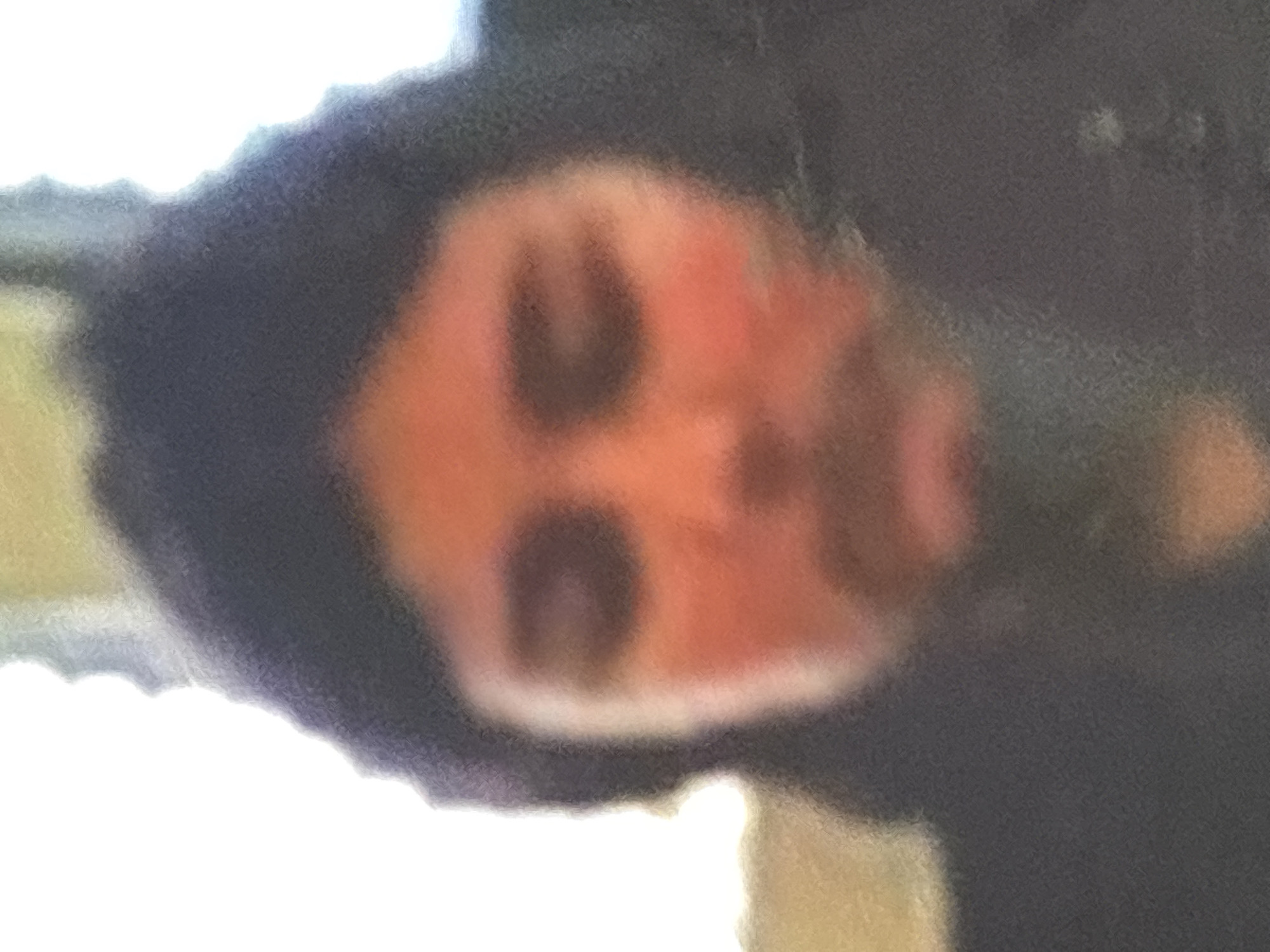 The image is a blurry, close-up, head-and-shoulders shot of a person. The subject appears to have dark hair and is wearing a dark jacket or hooded garment. The lighting is bright, suggesting an outdoor setting or a well-lit indoor space. The background is out of focus and appears to be a lighter color, possibly a wall or sky. No specific location in the city of Orihuela, Spain can be identified from this image. The subject's facial features are indistinct due to the blurriness.