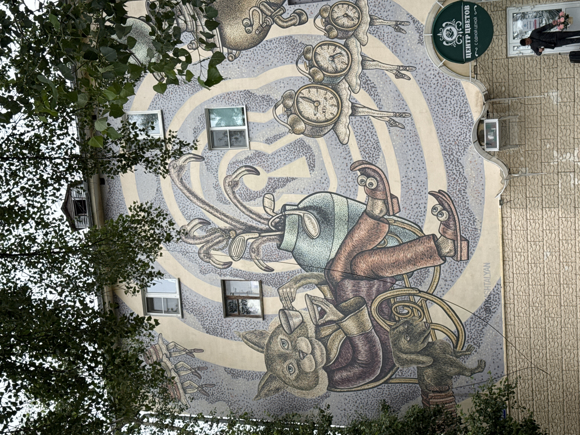The image depicts a large mural on the side of a multi-story building in Nizhny Novgorod, Russia. The mural features a whimsical and surreal scene with anthropomorphic characters. A cat, dressed in a robe and seated in a rocking chair, is holding teacups. Beside the cat is a dachshund. In the background, there are several alarm clocks on ballet dancer legs, and a vase filled with golf clubs. The building has several windows, and there is foliage from trees obscuring parts of the mural. Below the mural, on the ground floor, there is a storefront with a sign reading "ЦЕНТР ЦВЕТОВ" (Flower Center). A man is standing outside the storefront, holding a bouquet of flowers. The building's facade is made of brickwork.
