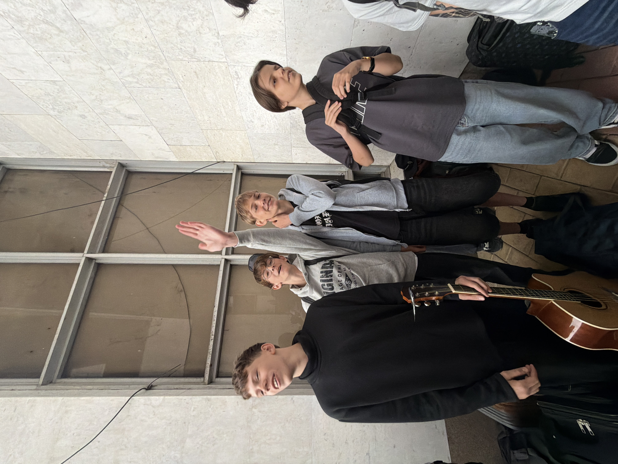 A group of four young people stand close together in a public space, likely a covered walkway or building exterior, in an indeterminate location. The space is characterized by large windows with metal frames and a light-colored tiled floor. One person wears a black long-sleeved shirt and holds an acoustic guitar. Another, wearing a gray sweatshirt, raises one arm. A third wears a gray hoodie and a fourth is dressed in gray with a black backpack. The group's arrangement appears informal, possibly suggesting a casual gathering. The specific context and purpose of the group are unclear. The location of Birobidzhan, Russia, is not apparent from the image.