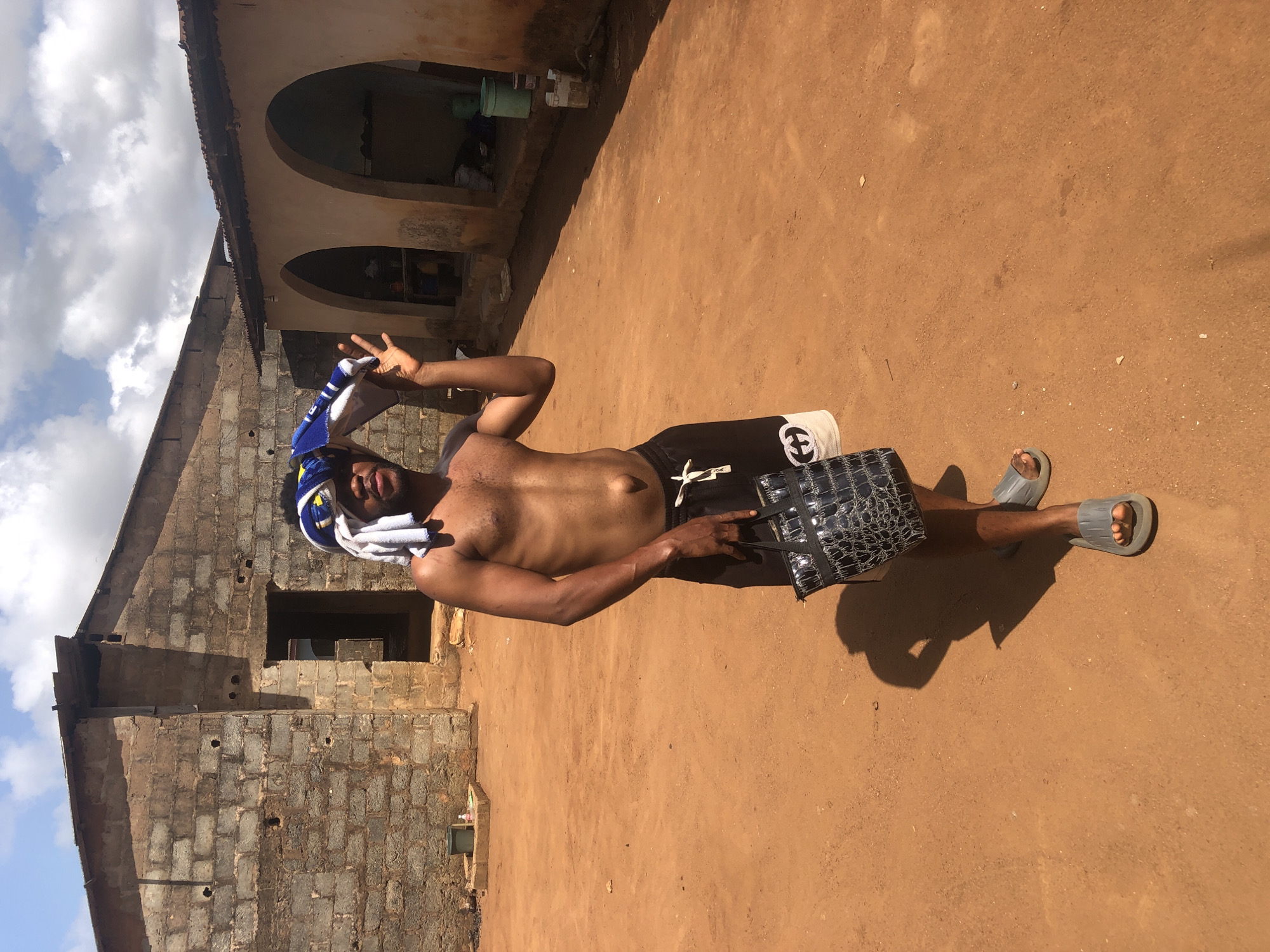 A topless man with a towel on his head stands on dirt ground in Unknown, Unknown. He is wearing shorts and sandals, holding a dark-colored bag. The background includes a brick building and another structure with arches. The sky with some clouds is visible in the upper left corner.